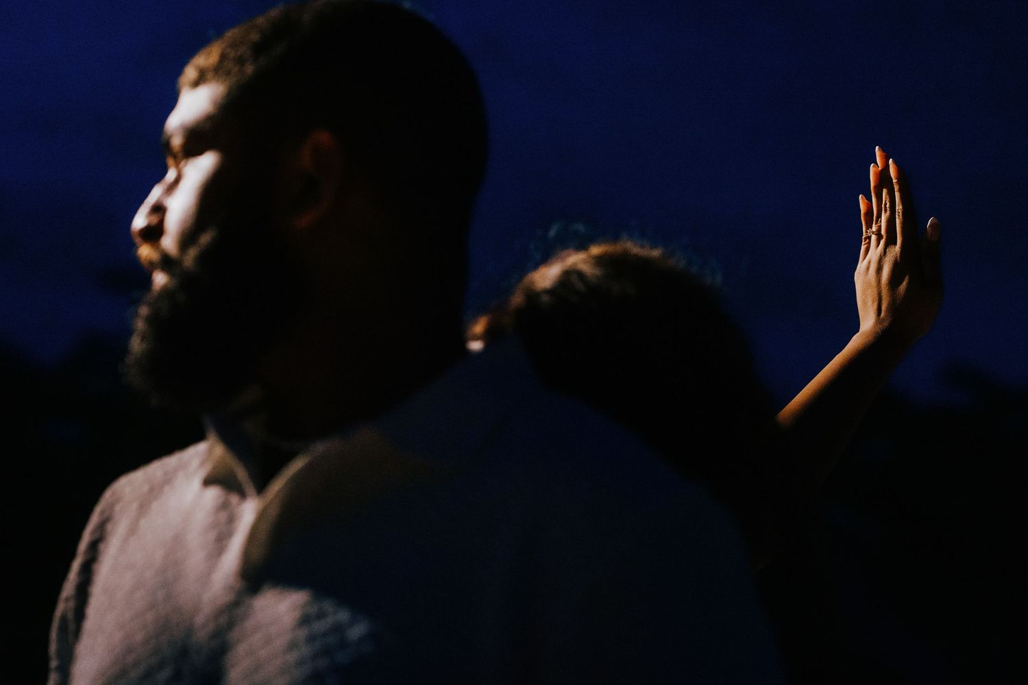 Silhouetted figures stand against a dark background with dramatic lighting and raised hands creating a moody atmosphere.