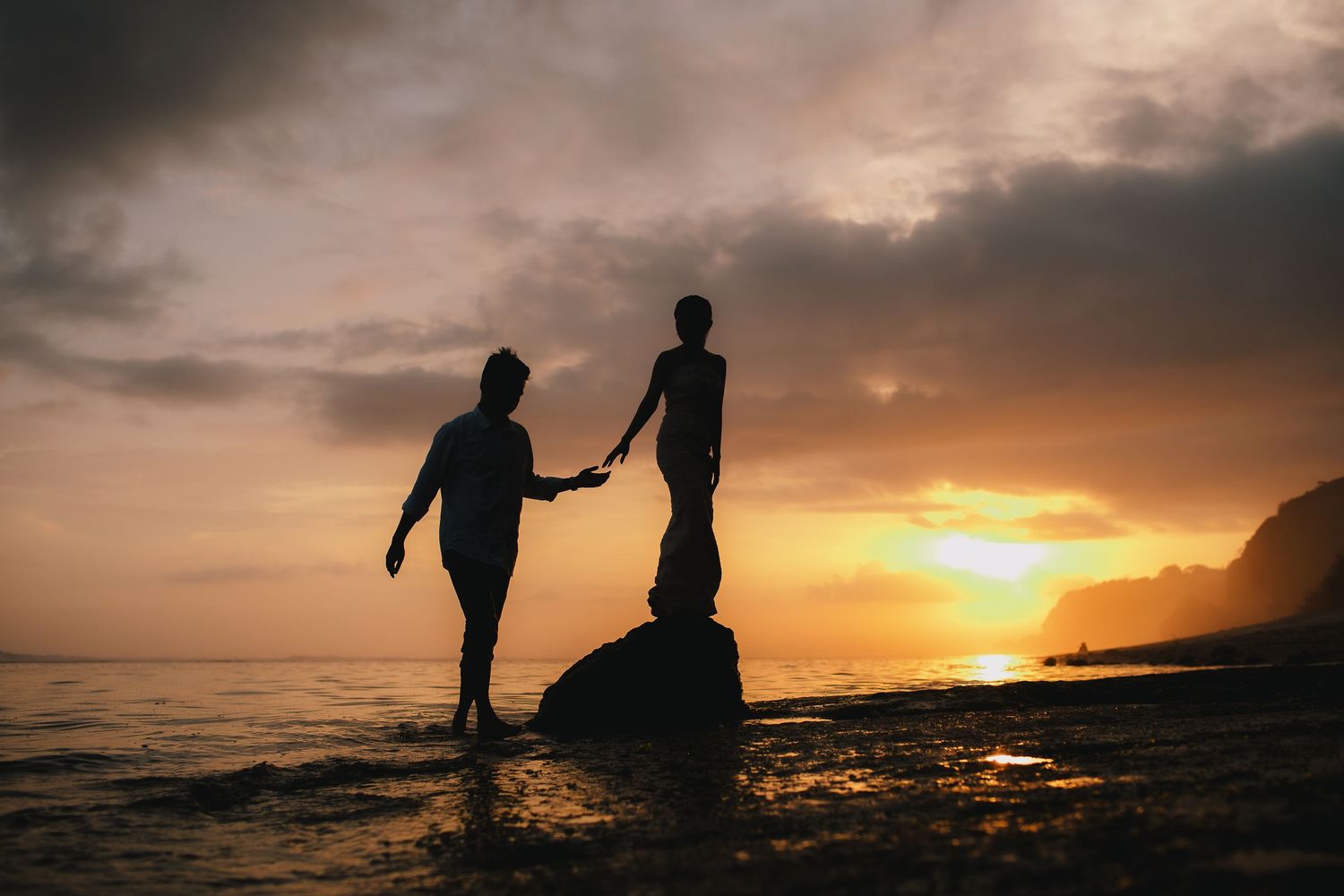 Silhouettes walking along beach shore during dramatic orange sunset.