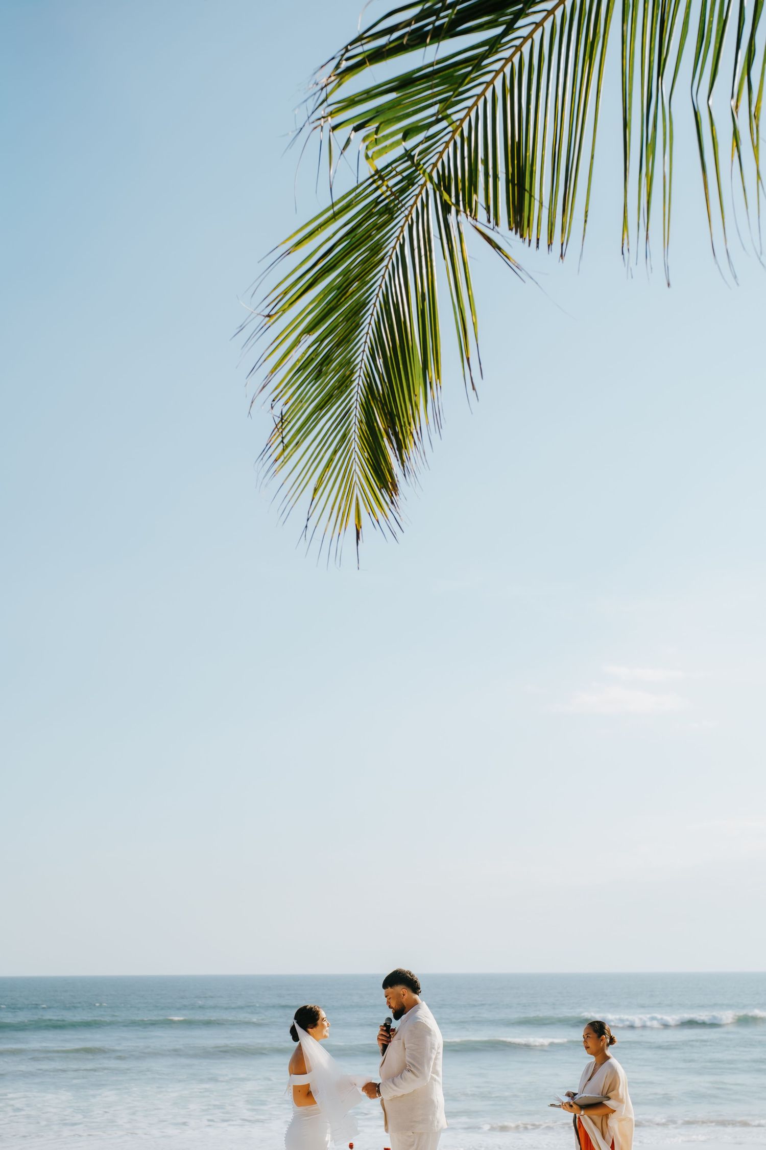 Couple exchanges vows on a tropical beach with palm fronds overhead and ocean waves in the background.