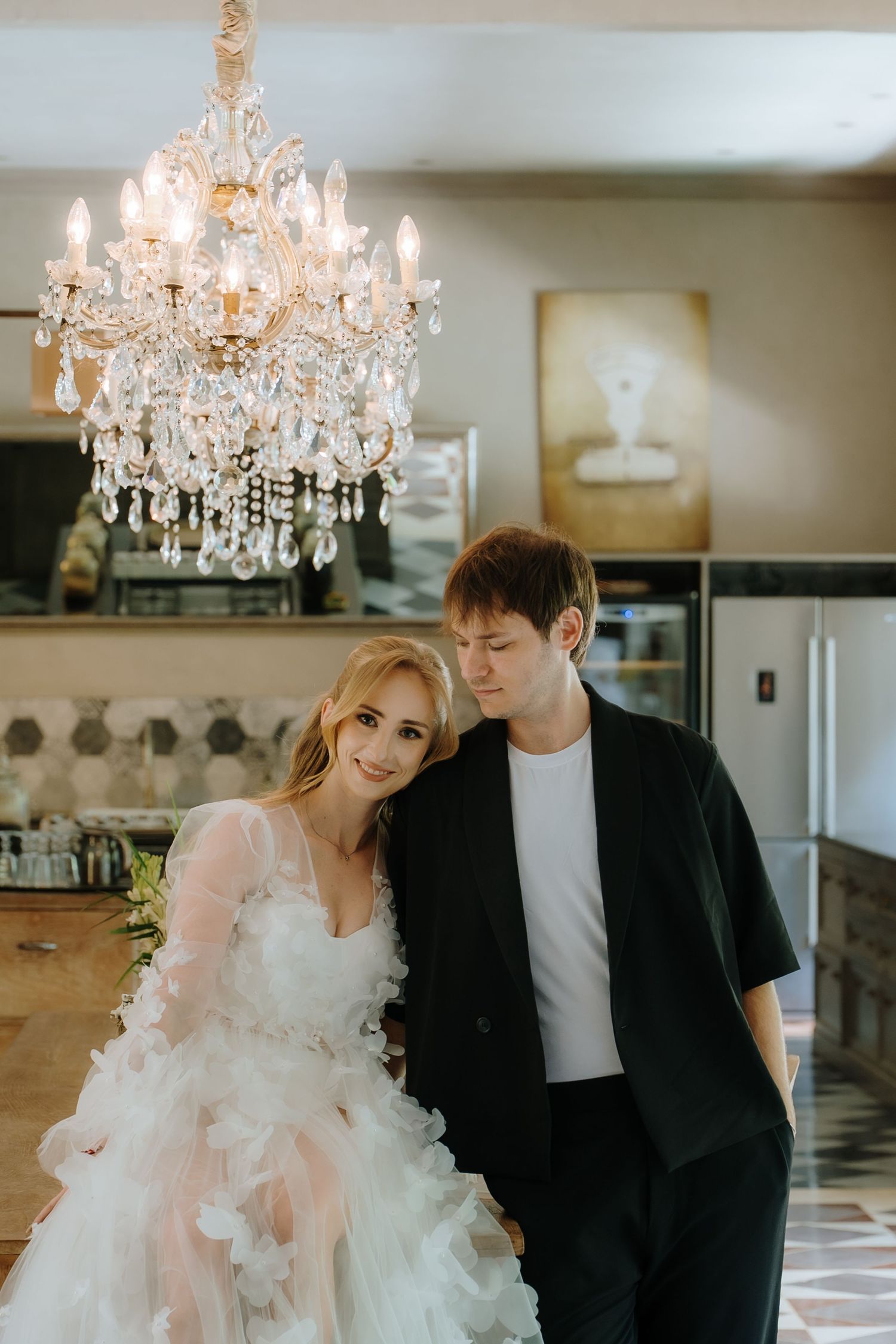 Bride in ruffled white gown and groom in black suit pose under crystal chandelier in elegant venue.
