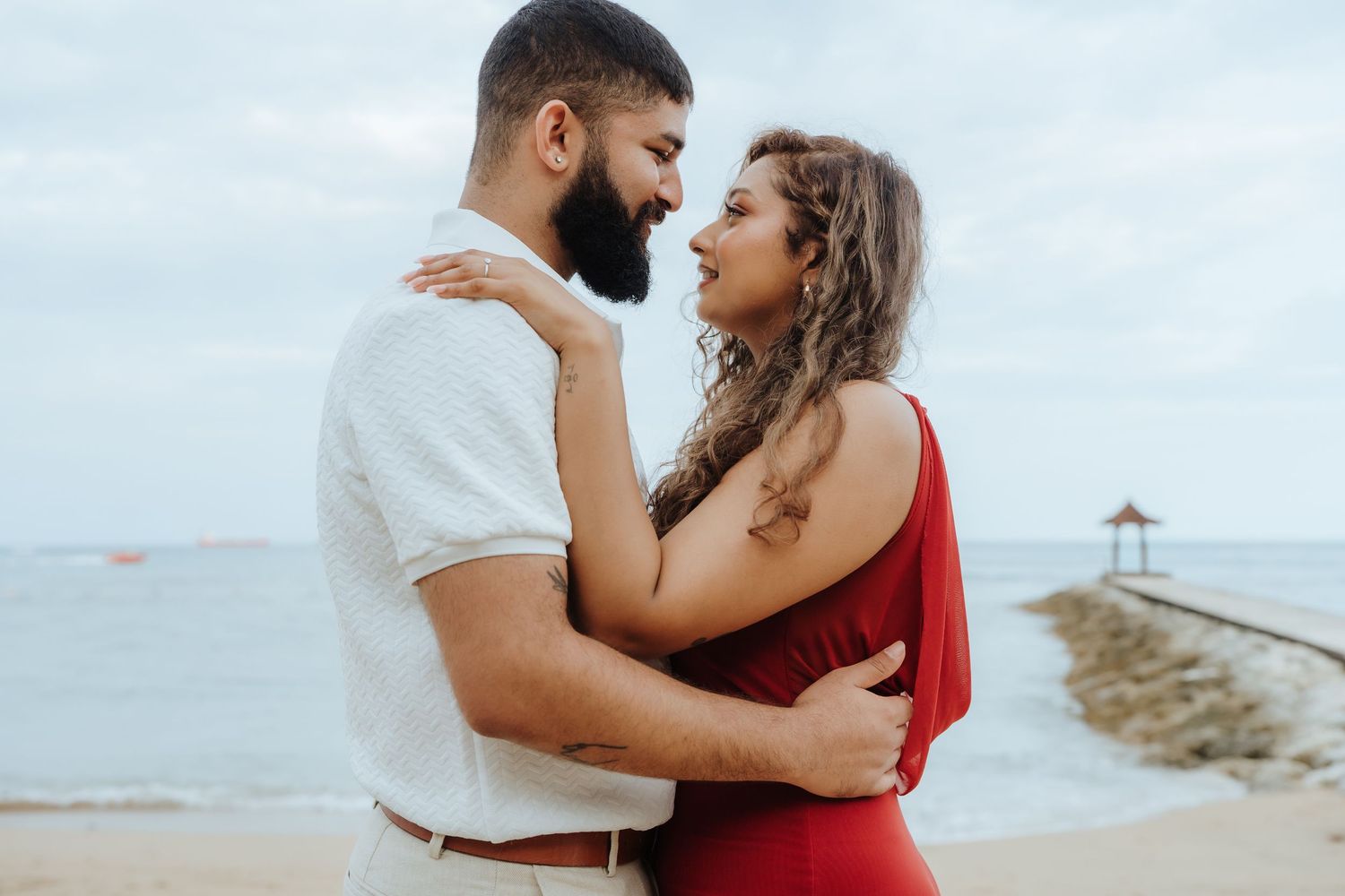 A couple in red and white outfits shares a tender moment by the ocean.