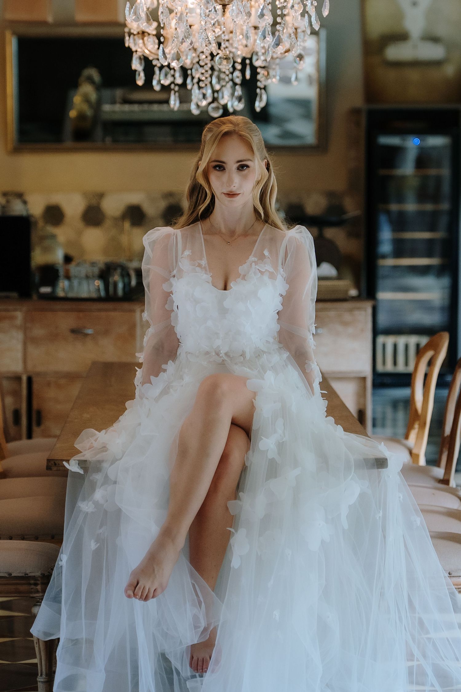 Bride in a white floral tulle gown sits elegantly under a crystal chandelier in a vintage-styled venue.