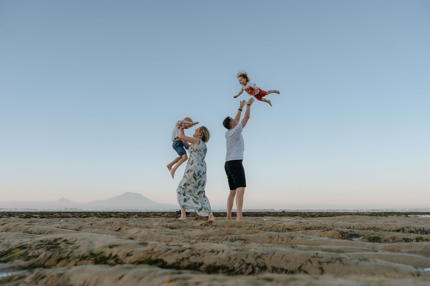 Family of four playing on a sandy beach at sunset, parents joyfully lifting two young children into the clear blue sky.