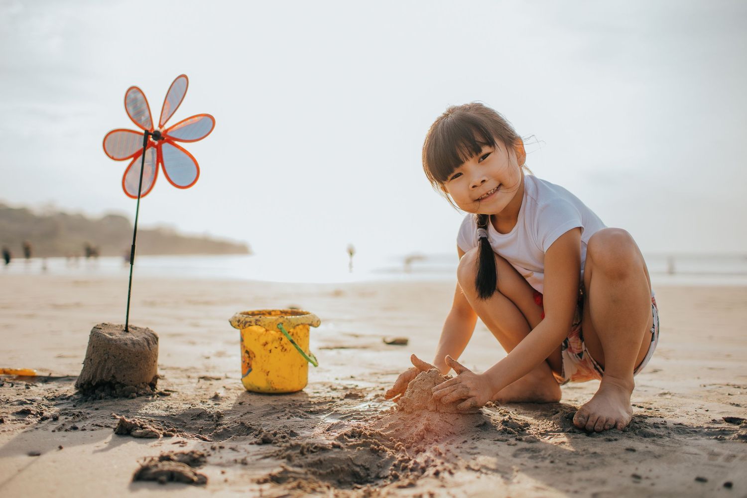 Child digging in sand next to pink flower decoration and yellow bucket at the beach.