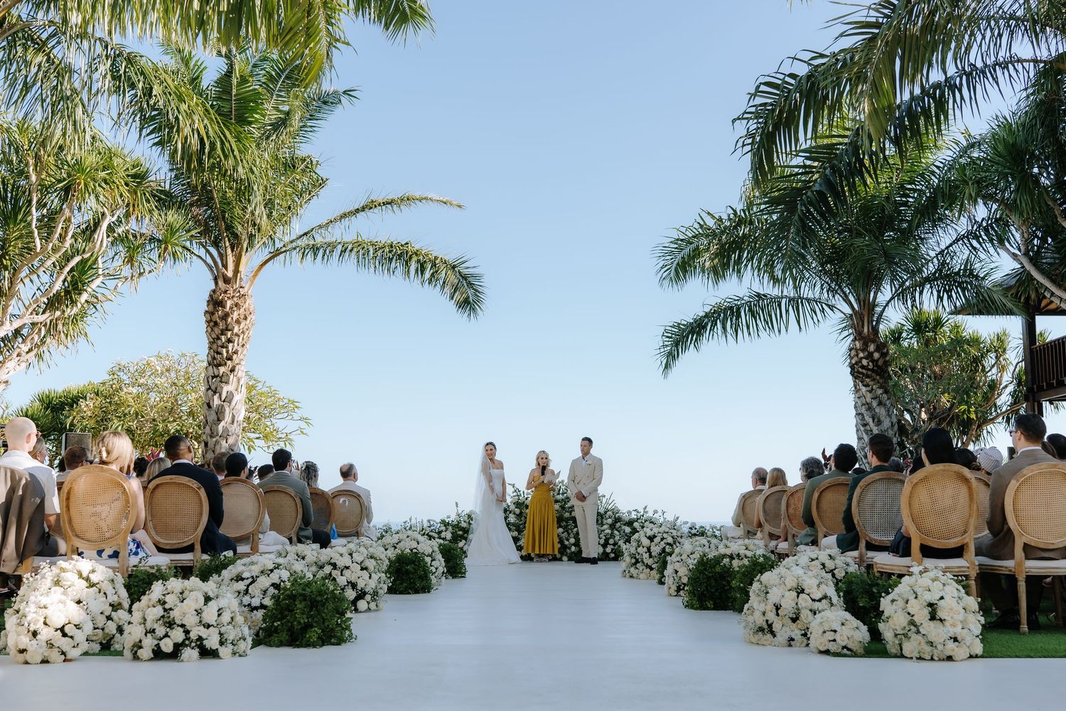 Outdoor wedding ceremony with white floral aisle, rattan chairs, palm trees, and ocean backdrop under clear blue sky.