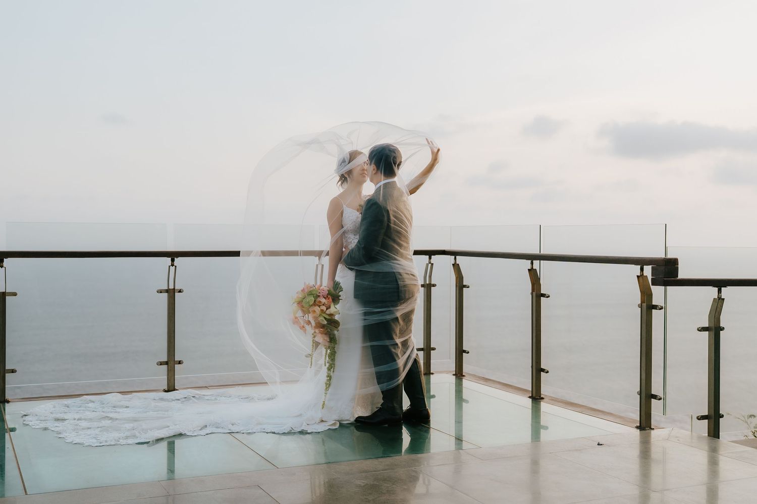 A bride and groom stand on an ocean-view terrace, veil flowing in the breeze, sharing a romantic wedding moment.