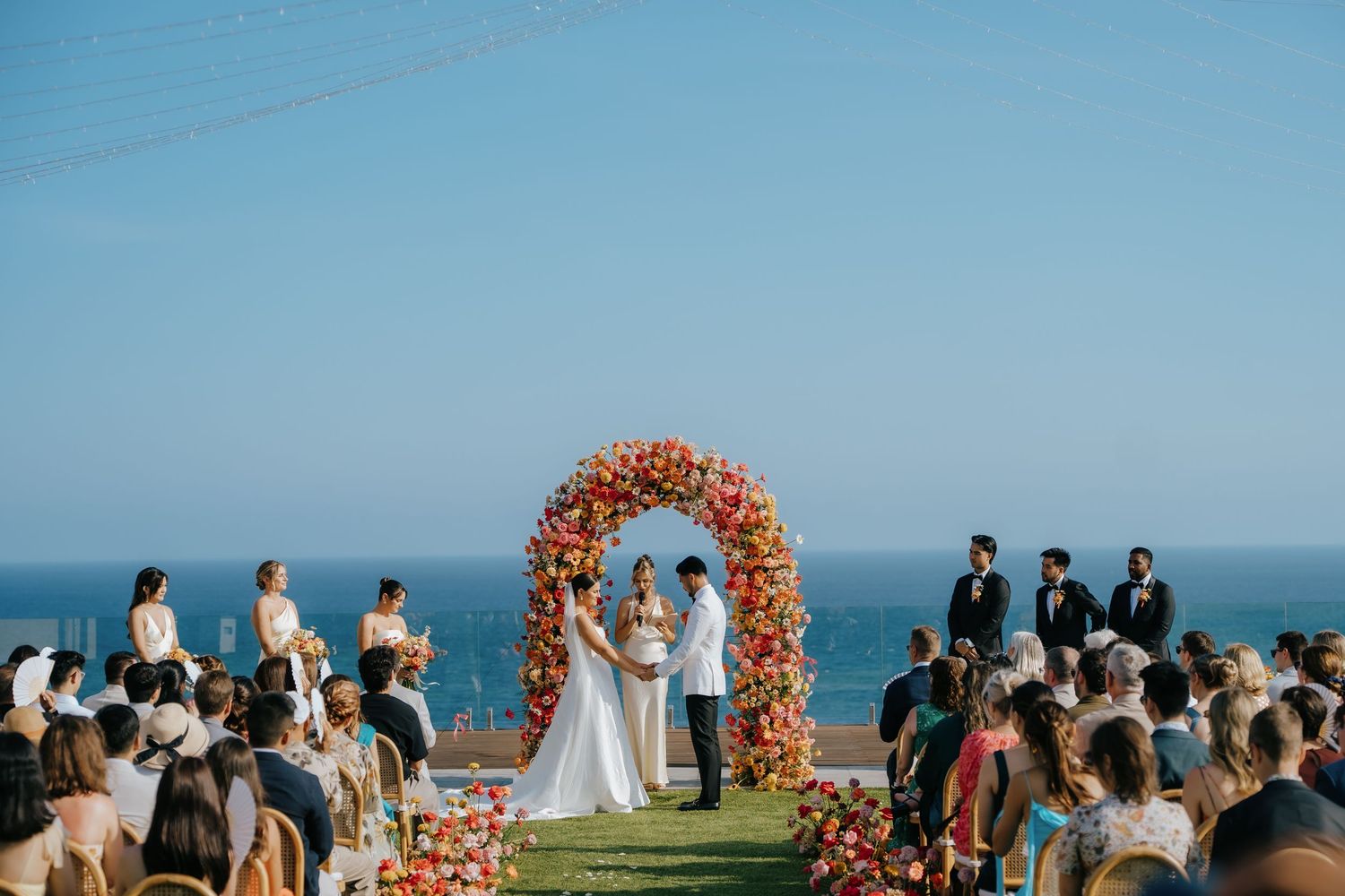 Outdoor wedding ceremony with floral arch overlooking the ocean, bride and groom exchanging vows before seated guests.