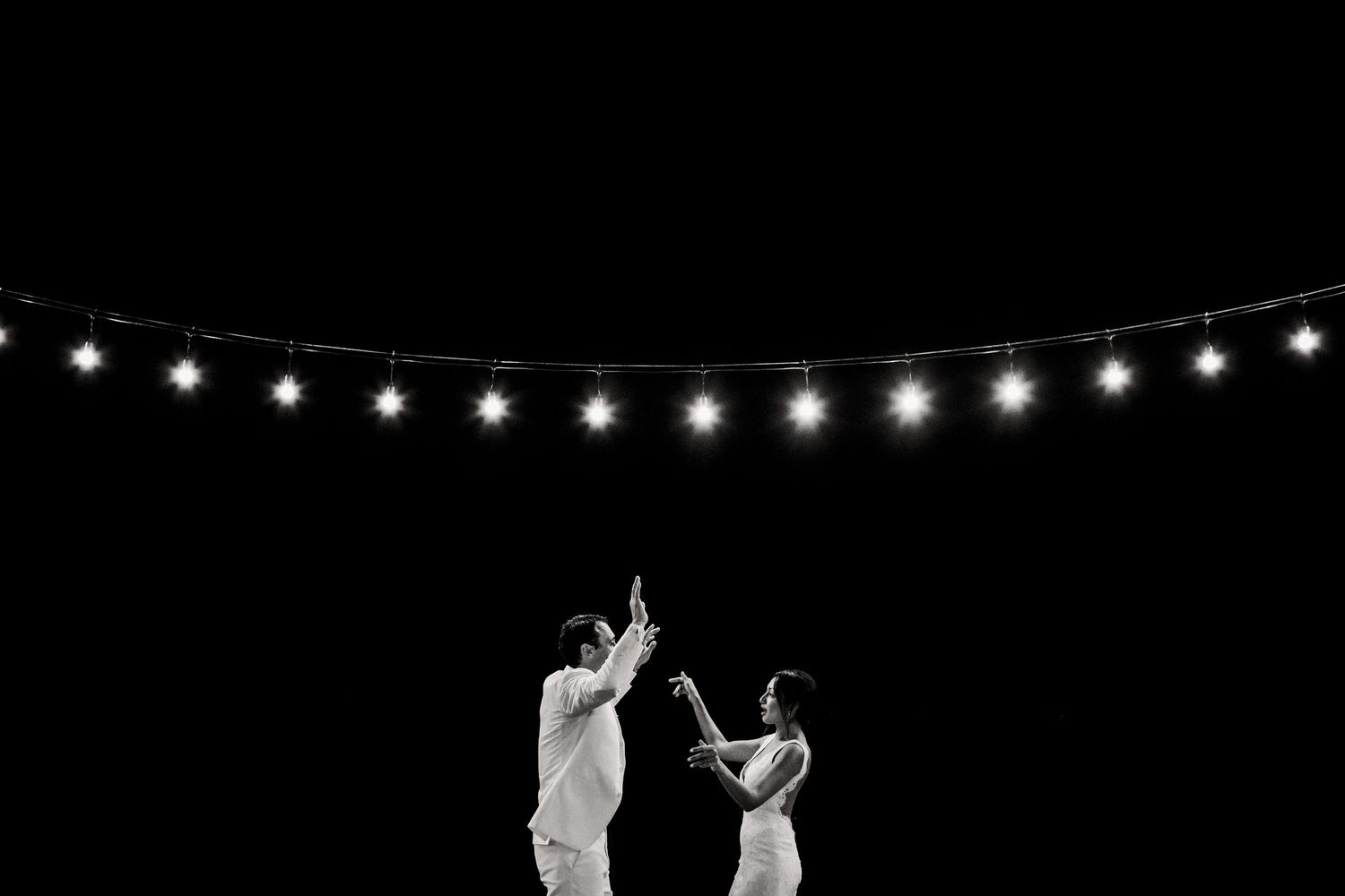 Black and white photo of a couple dancing under string lights against a dark night sky.