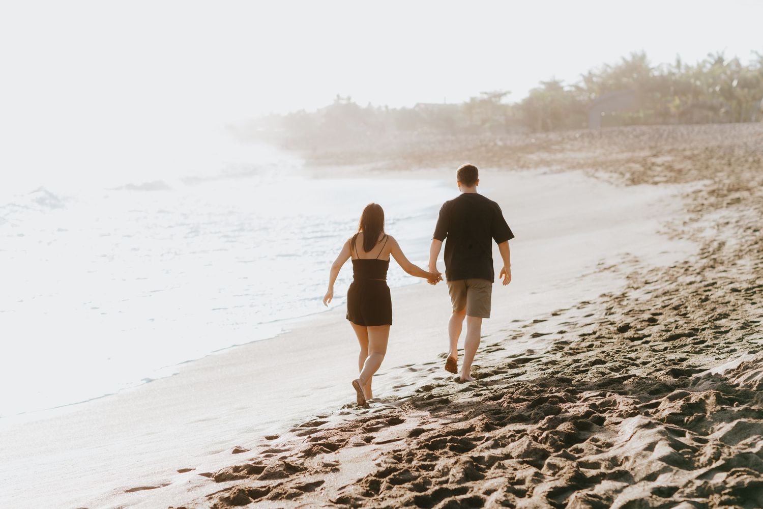 A couple walks along a misty beach shoreline during sunset with palm trees in the background.