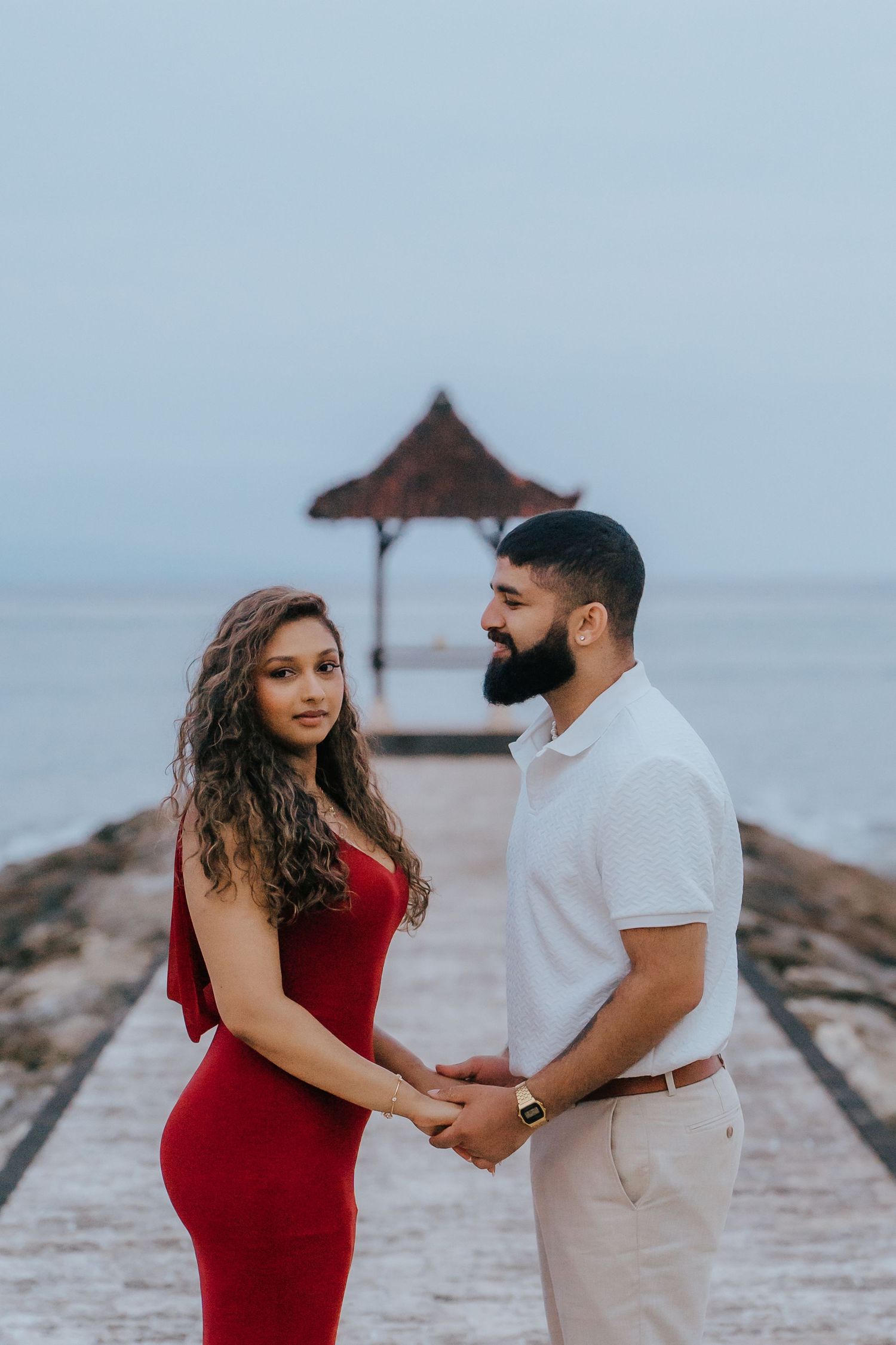 A couple stands together on a rocky beach, with the woman wearing a red dress and the man in white clothing.