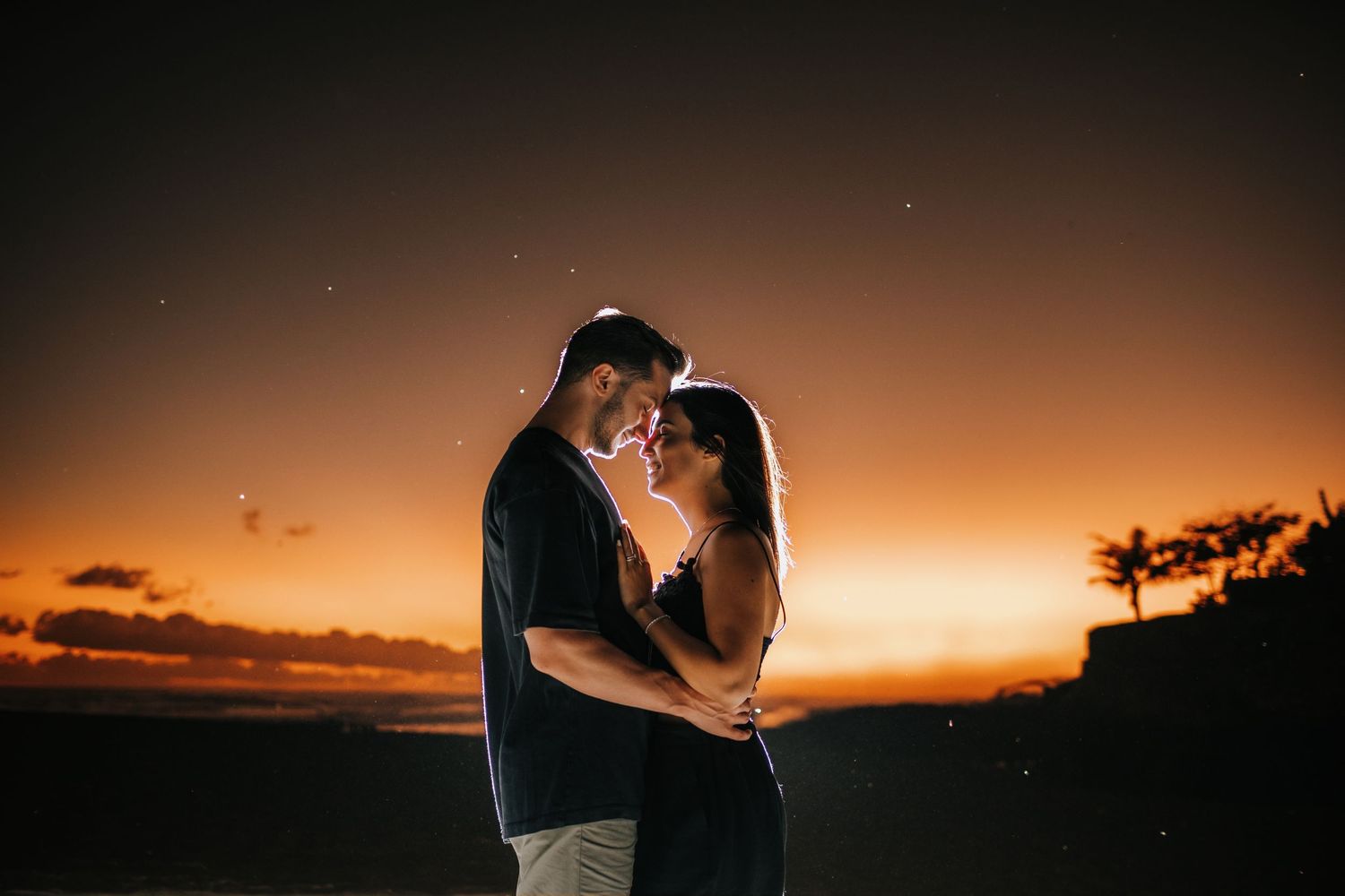 Couple sharing intimate moment against glowing orange sunset sky.