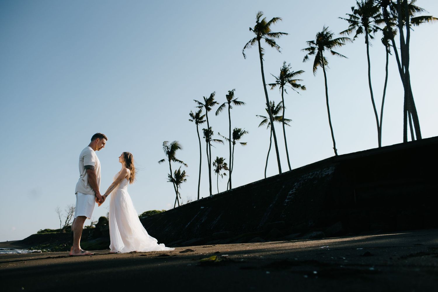 A couple stands on a dark beach with tall palm trees silhouetted against a blue sky in Hawaii.