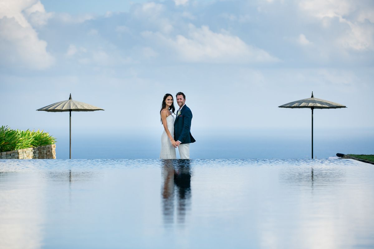 Wedding couple standing between beach umbrellas at infinity pool overlooking ocean.