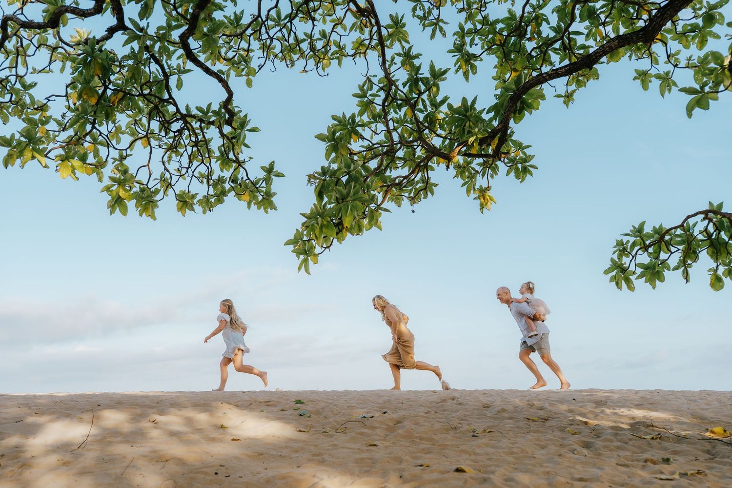 Three children playing and running on a sandy beach under lush green tree branches with a clear blue sky.