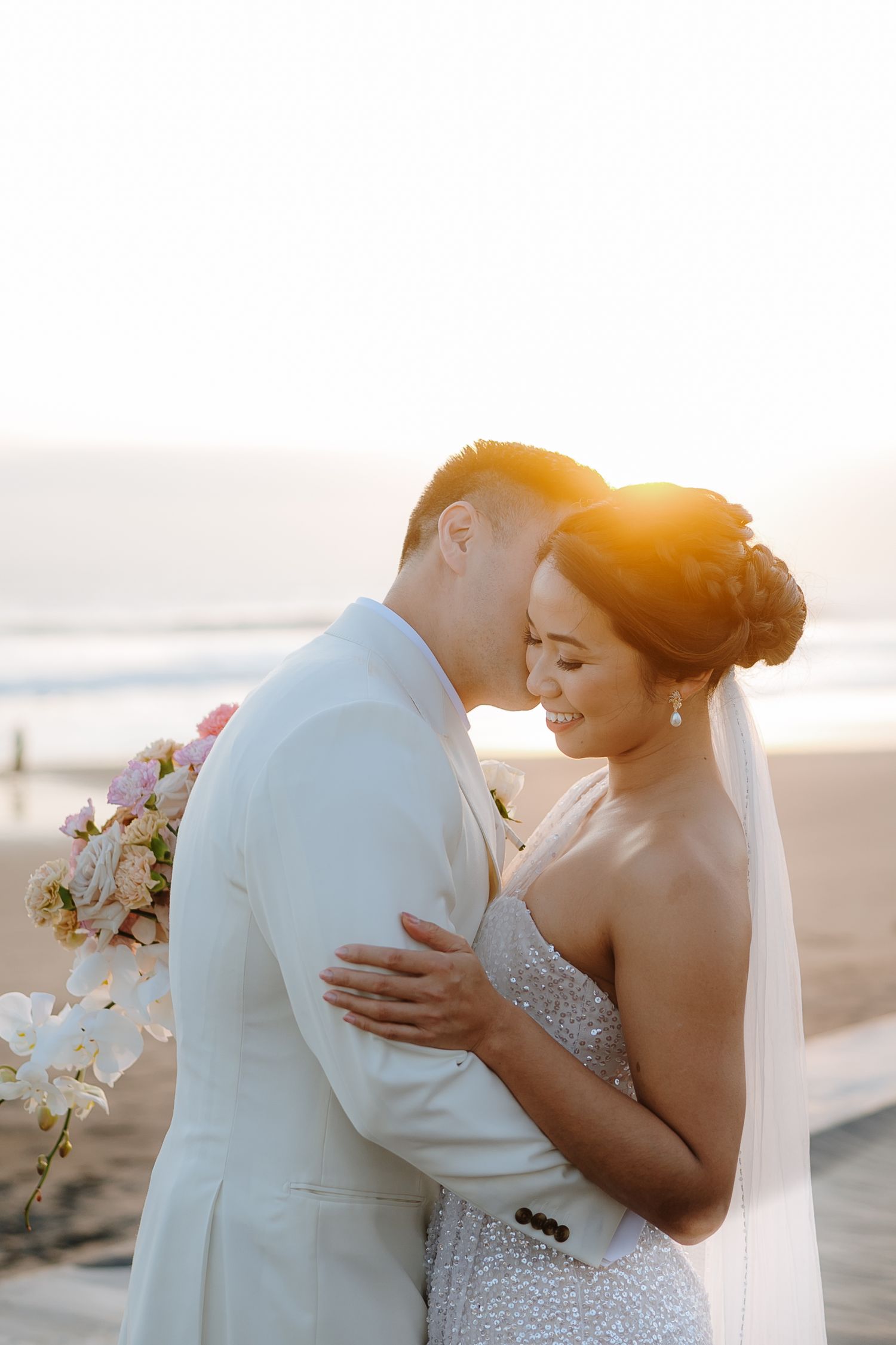 A bride and groom share an intimate moment at sunset on a beach, with golden light glowing behind them.