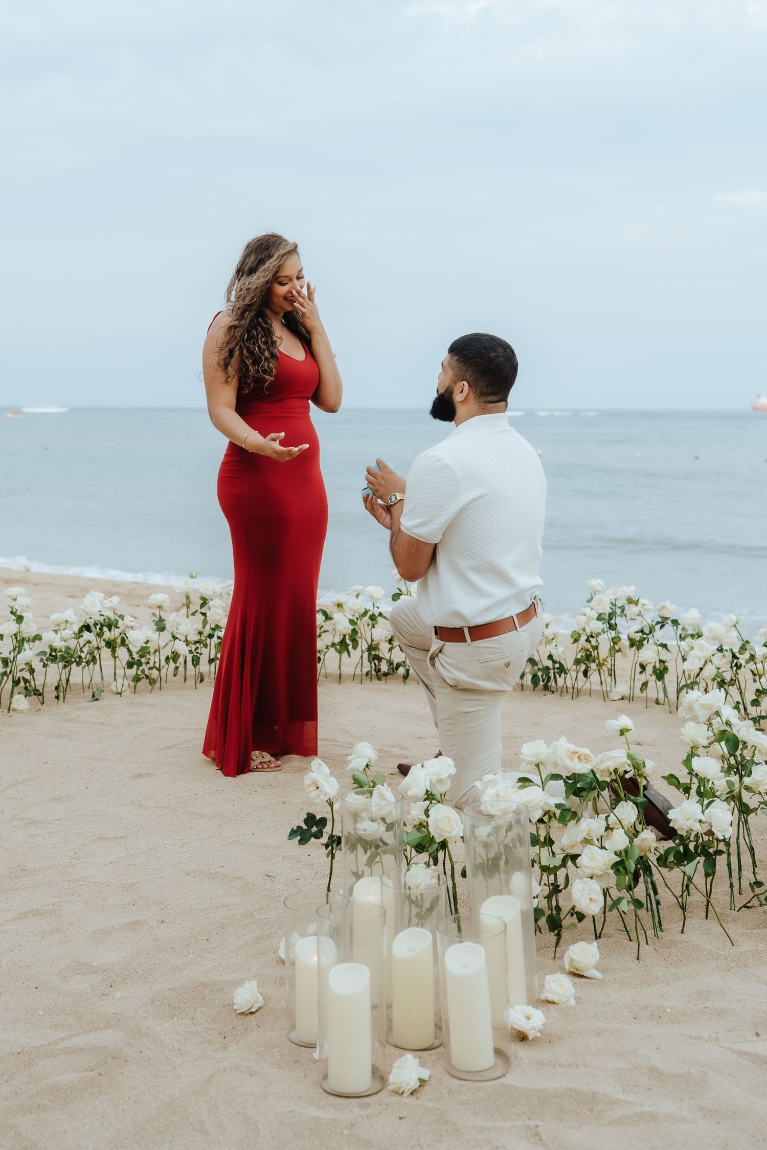 A romantic beach proposal scene with white candles and flowers arranged on the sand as waves roll in the background.