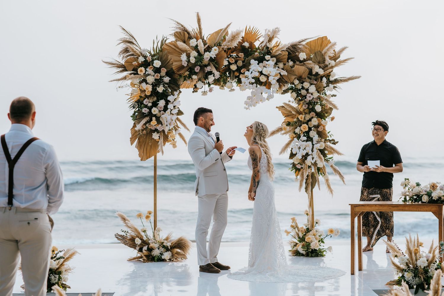 Couple exchanging vows at a boho beach wedding under a floral arch with dried pampas grass and white blooms.