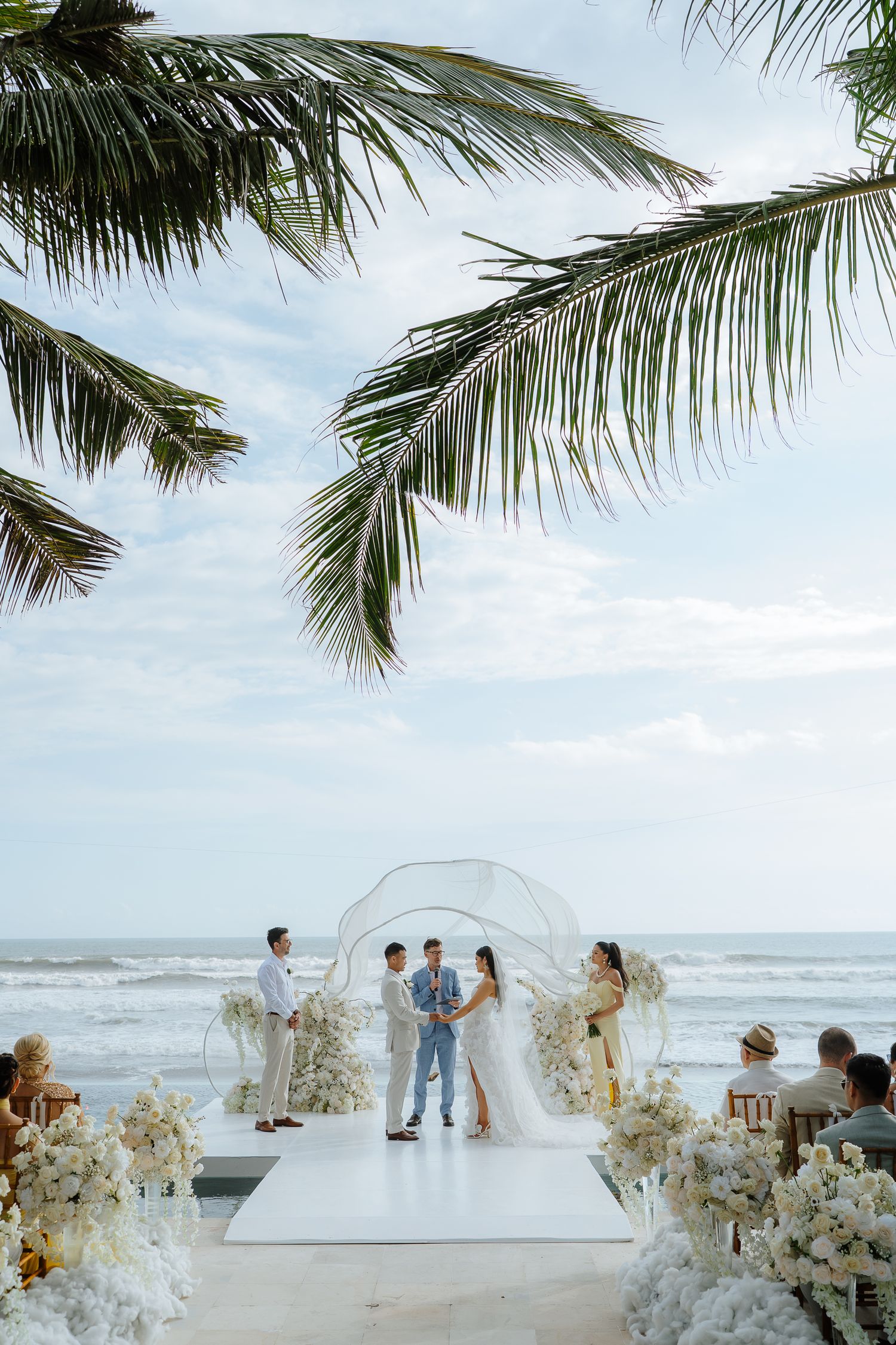 Couple exchanges vows at a beachside wedding ceremony with white floral arch, palm trees, and ocean backdrop.