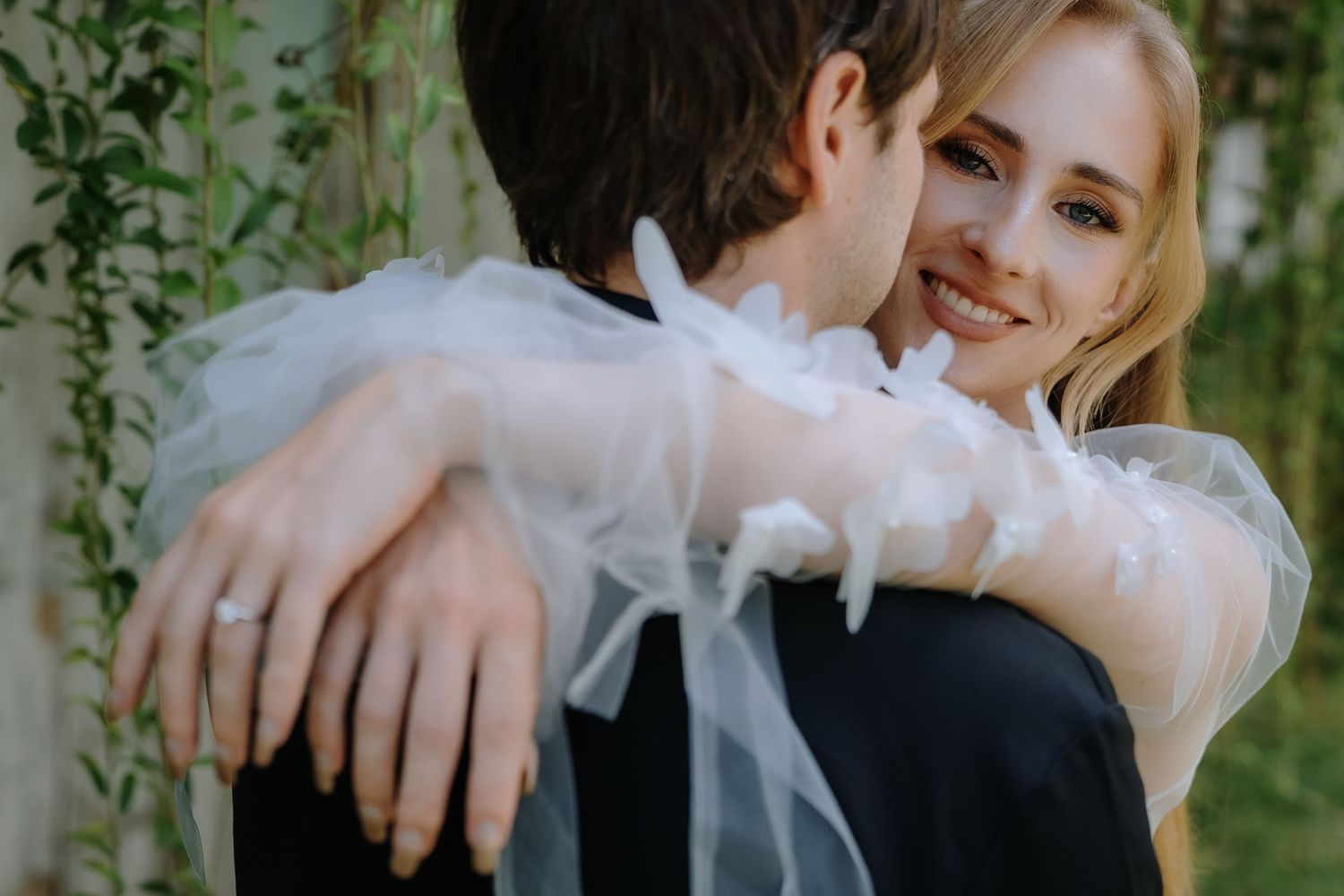 A smiling blonde bride in a white ruffled gown embraces her groom outdoors at a garden wedding ceremony.