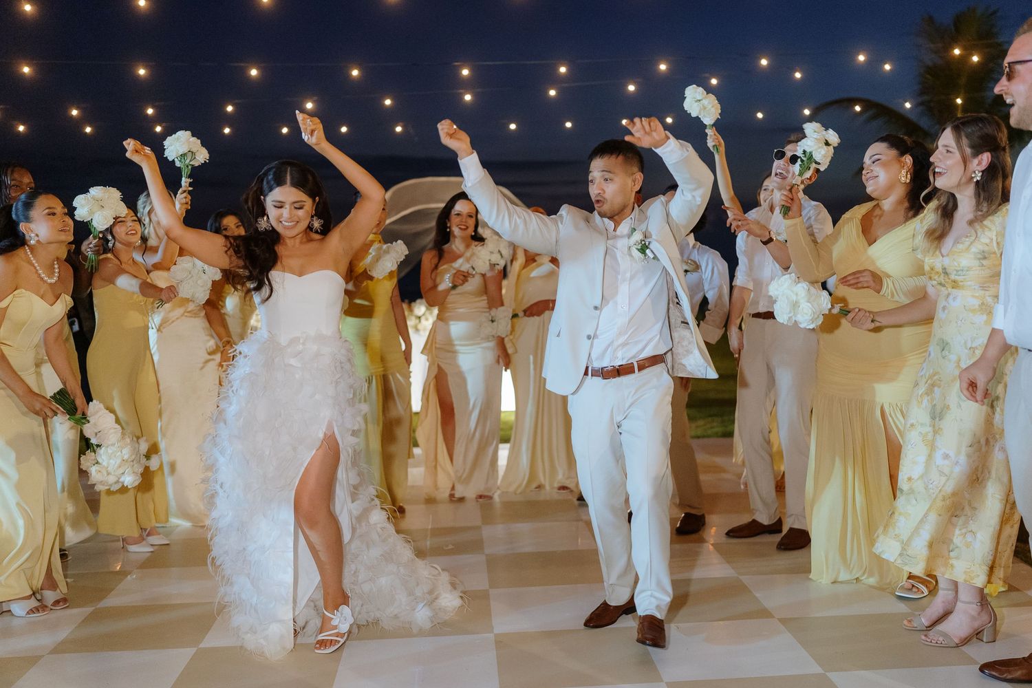 Bride and groom celebrate joyfully with bridesmaids in yellow gowns under string lights at an outdoor wedding reception.