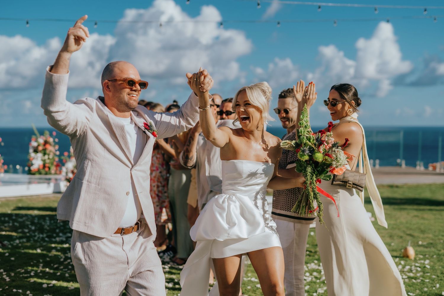 Joyful couple in white attire celebrating wedding ceremony outdoors near ocean, with guests throwing confetti.
