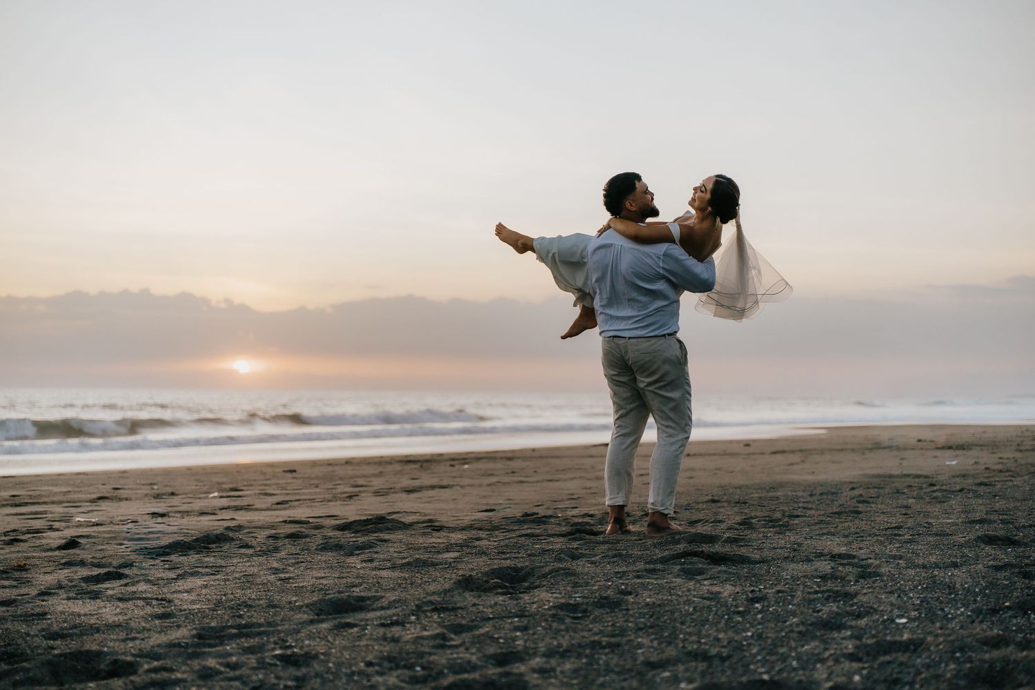 Couple dancing romantically on a beach at sunset, man lifting woman in white dress near ocean waves.