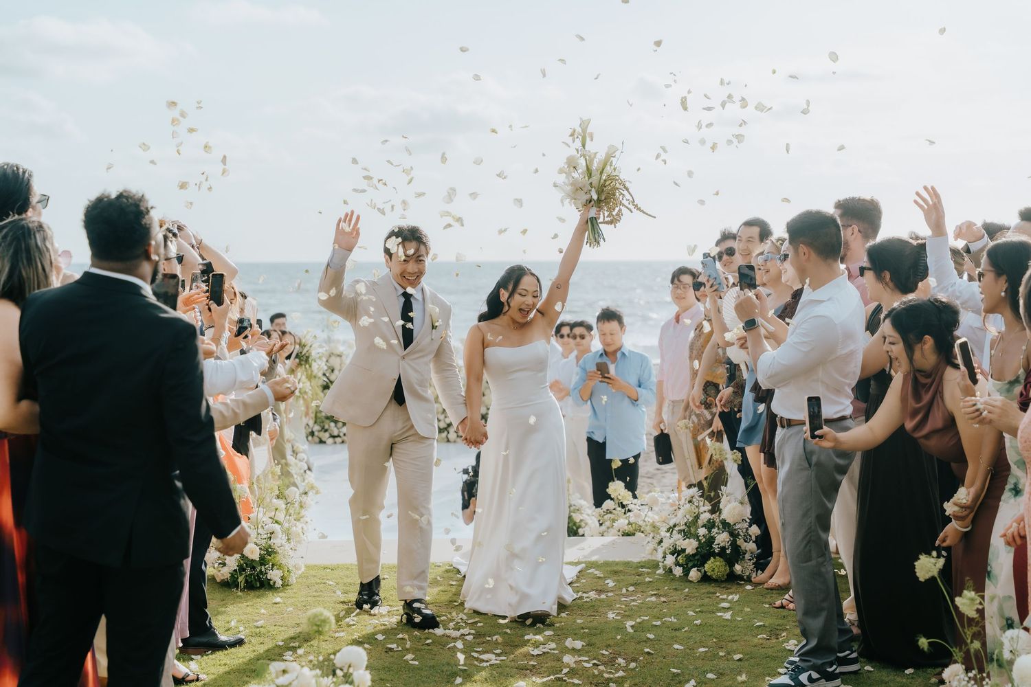 Joyful newlyweds walk down outdoor aisle as guests toss flower petals at beachside wedding ceremony.