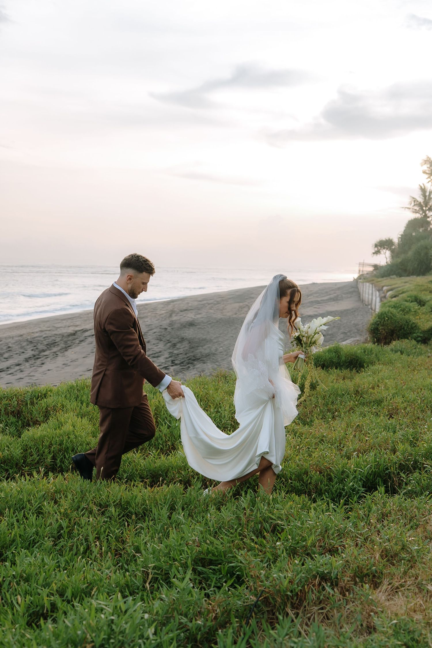 Bride and groom walking on grassy coastal bluff at sunset, groom in brown suit holding bride's white gown and veil.
