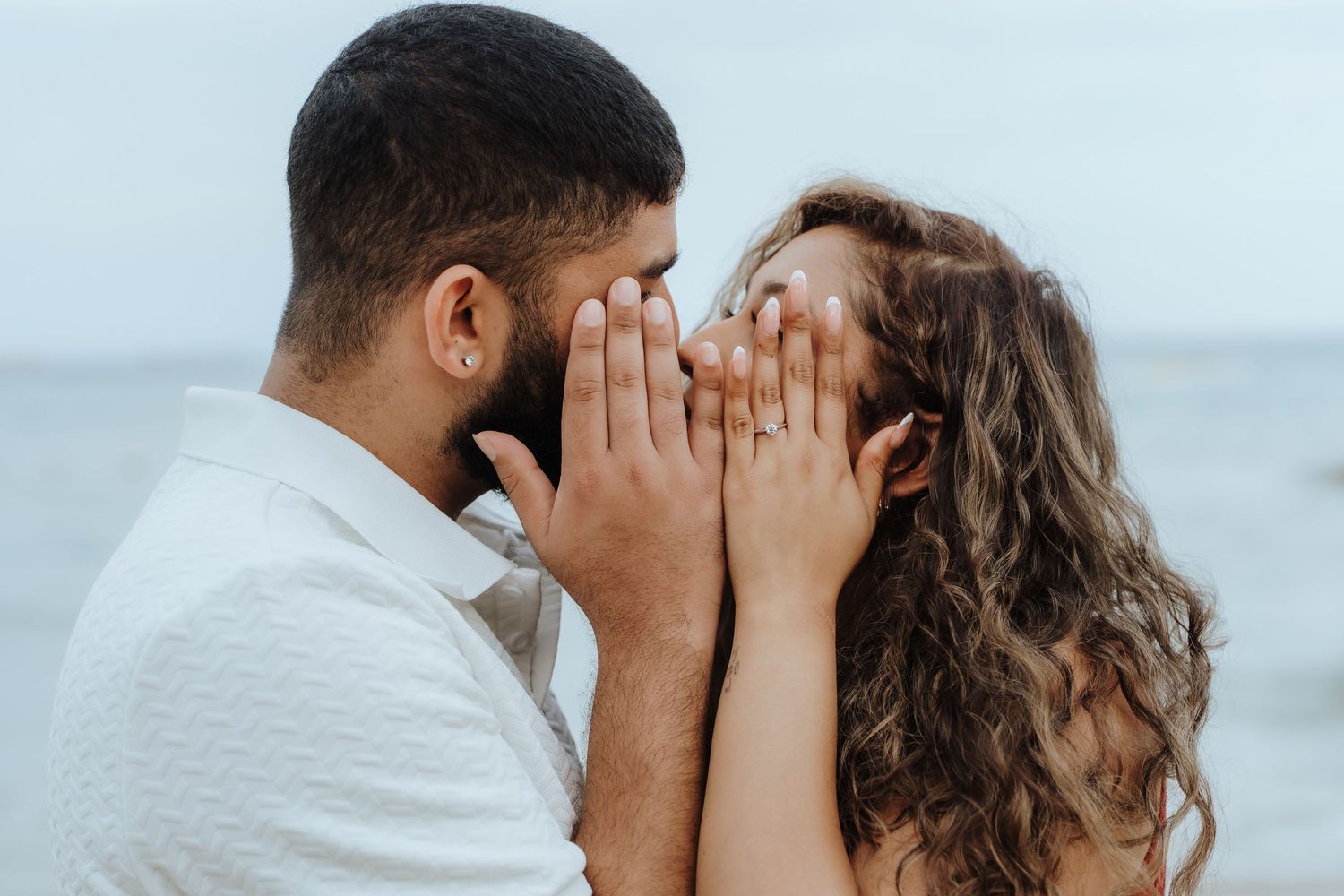 Couple shares intimate moment on beach with palm trees and cloudy sky in background.