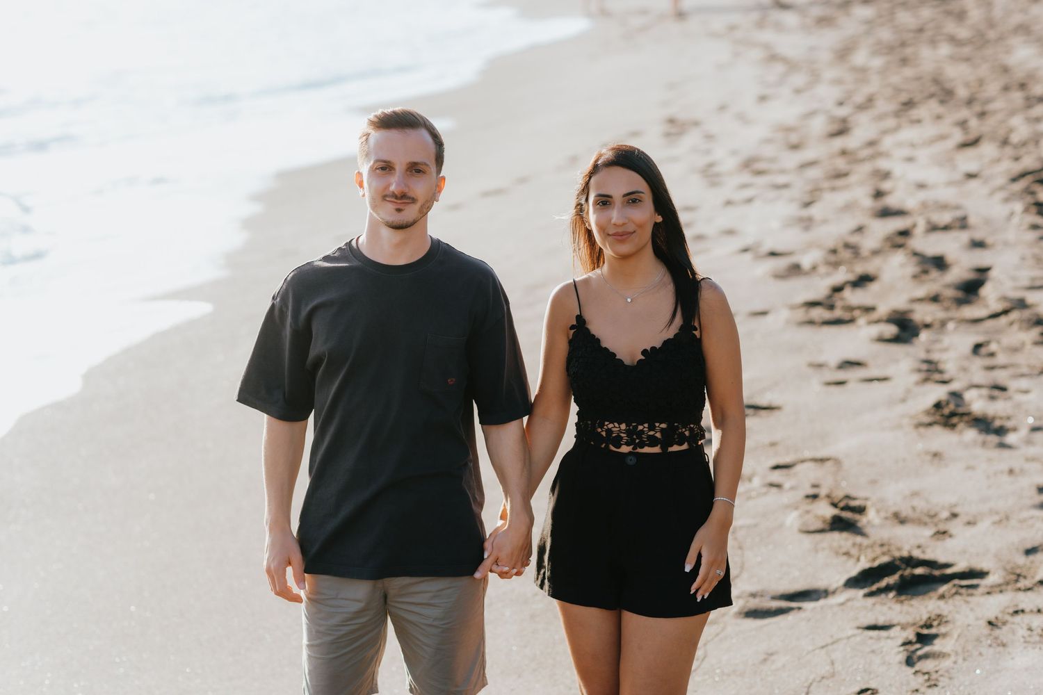 Couple walking together on sandy beach wearing casual black and gray outfits.