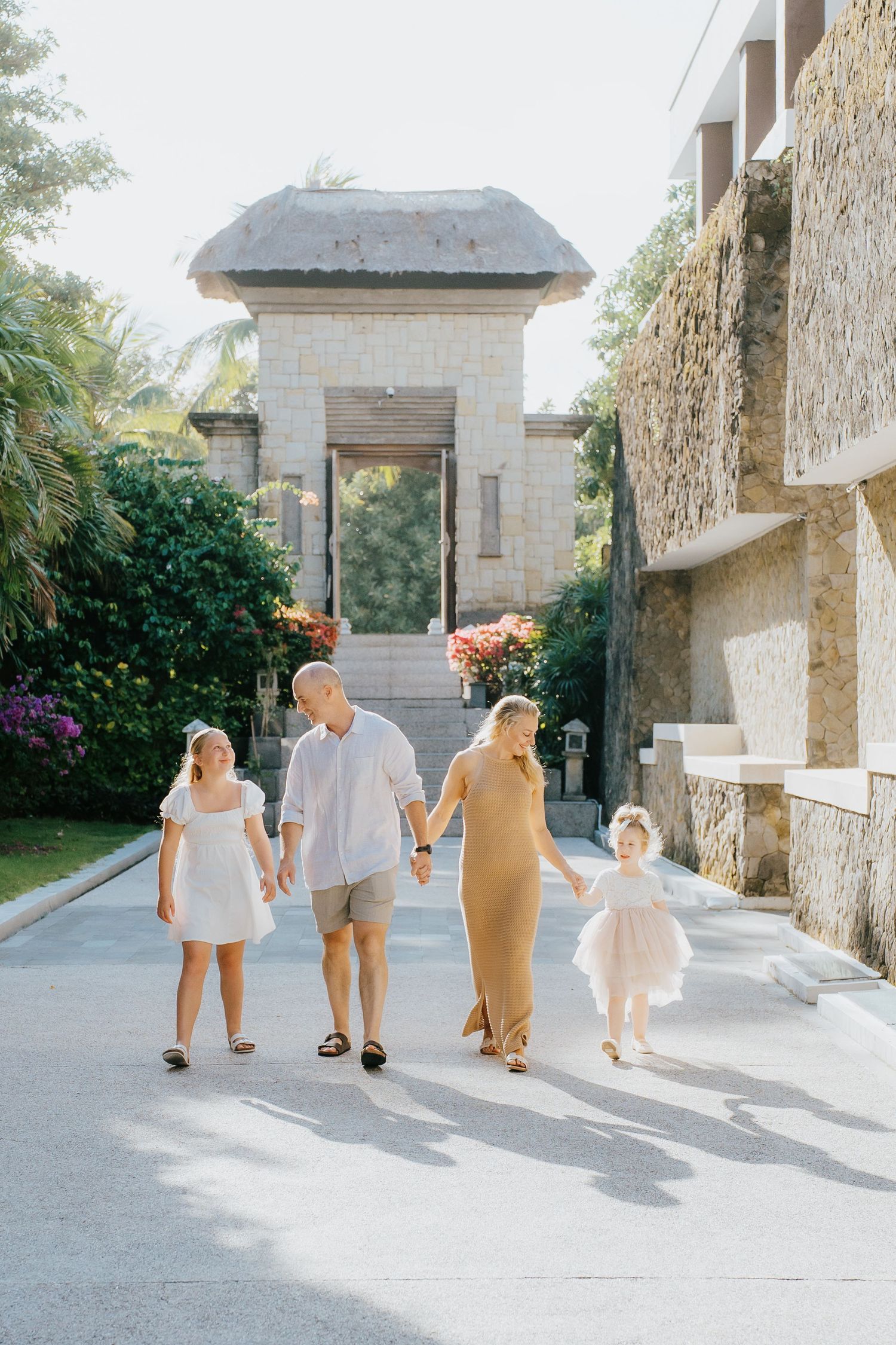 Family of four holding hands walking down a resort pathway surrounded by lush tropical gardens and stone architecture.