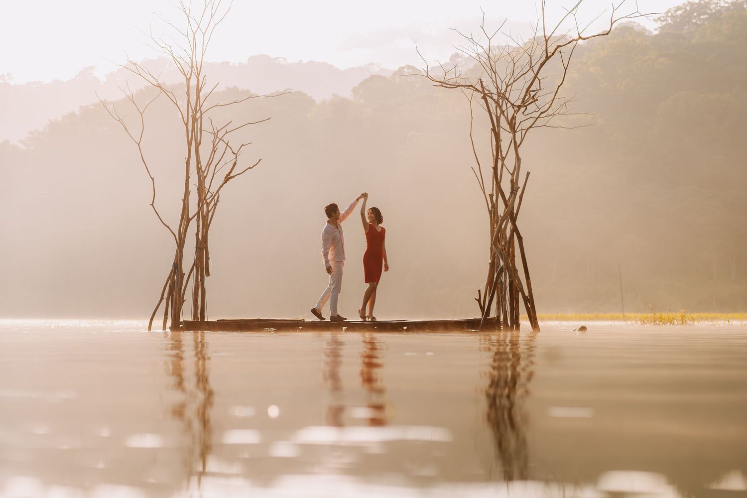 Couple dancing on a wooden platform over a misty lake surrounded by bare trees in golden morning light.