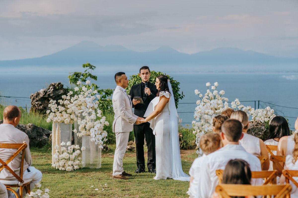 Outdoor wedding ceremony with couple exchanging vows, white floral arch, ocean and mountain views, guests seated in white attire.