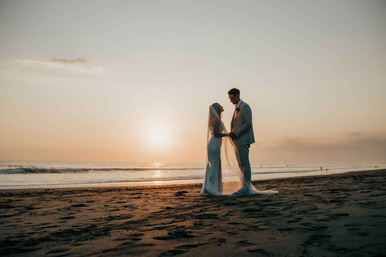 Silhouetted couple in wedding attire standing together on a beach at sunset with ocean waves in the background.