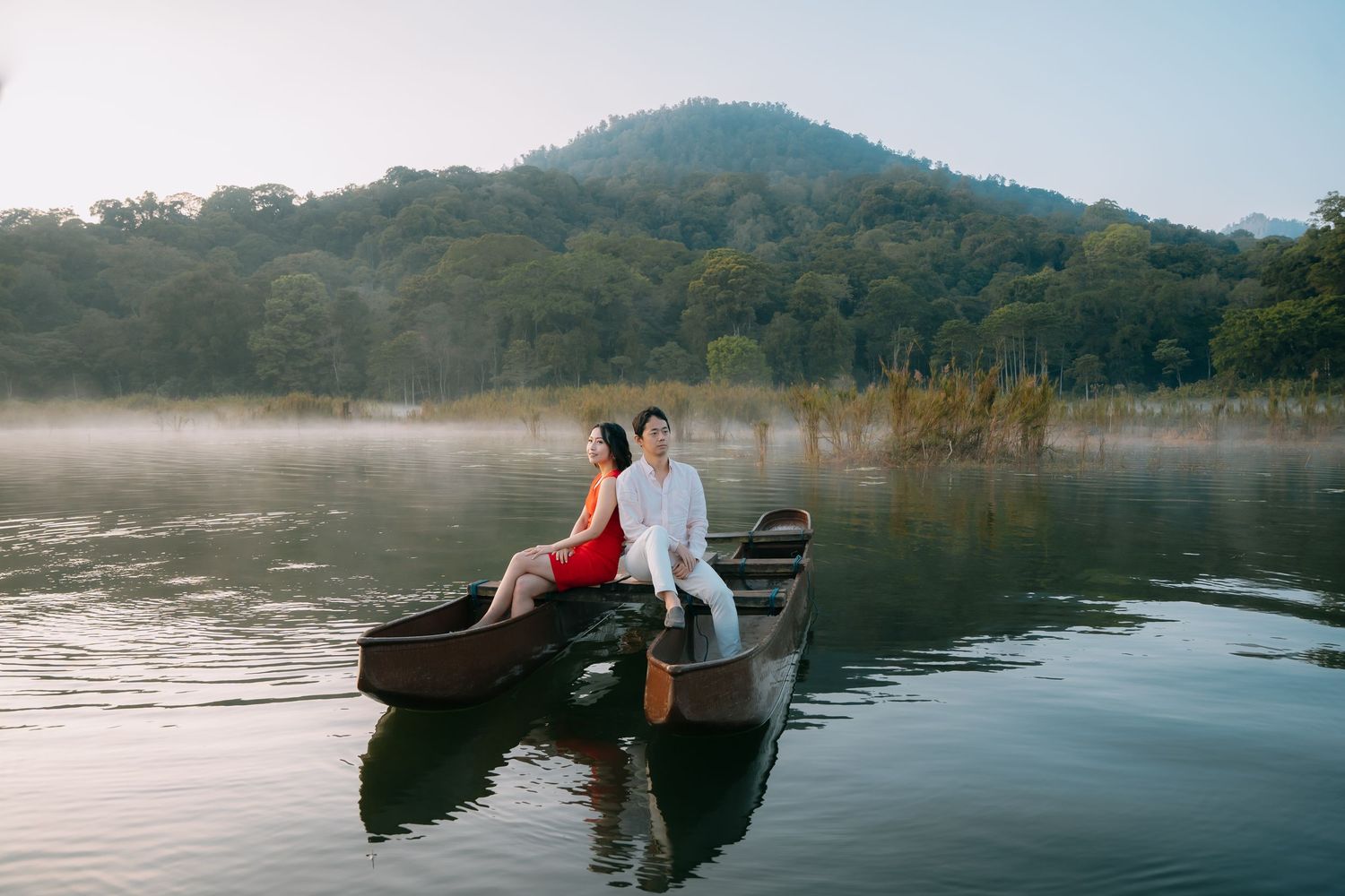 Couple in elegant attire sit in a wooden boat on a misty lake with mountain backdrop.