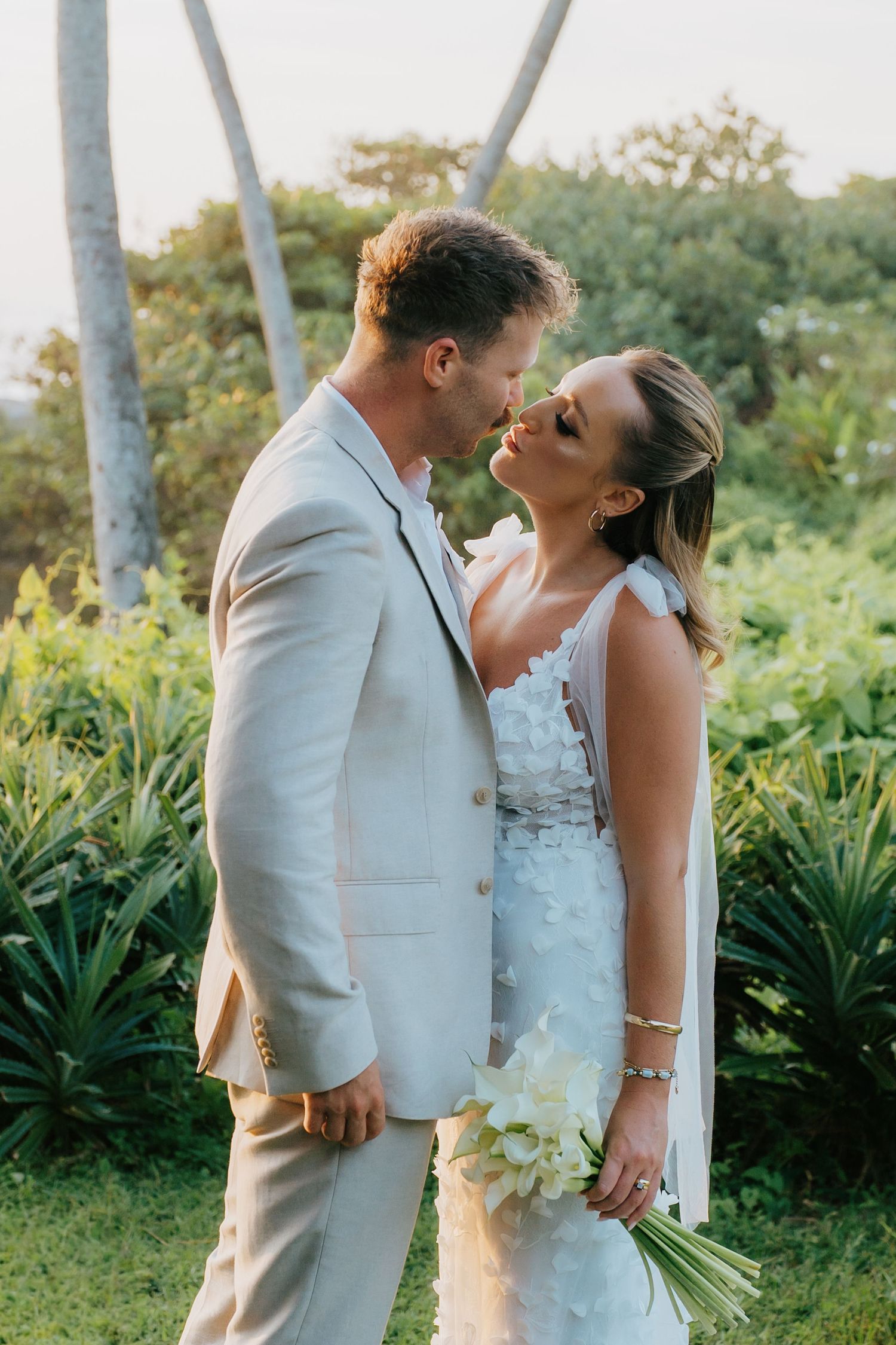 Bride and groom share a romantic moment outdoors, surrounded by lush tropical greenery, bride holding white flowers.