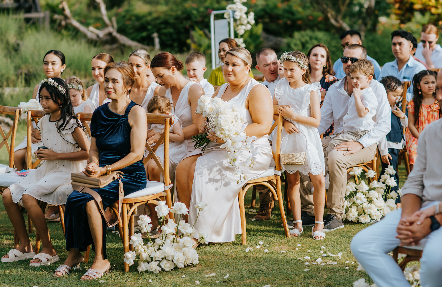 Bride holding white bouquet seated among wedding guests at an outdoor garden ceremony on a sunny day.