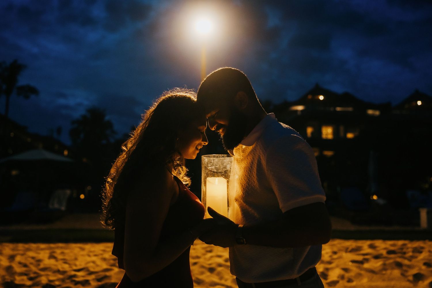 Silhouetted couple shares romantic moment on beach at sunset with glowing lantern creating warm ambiance.