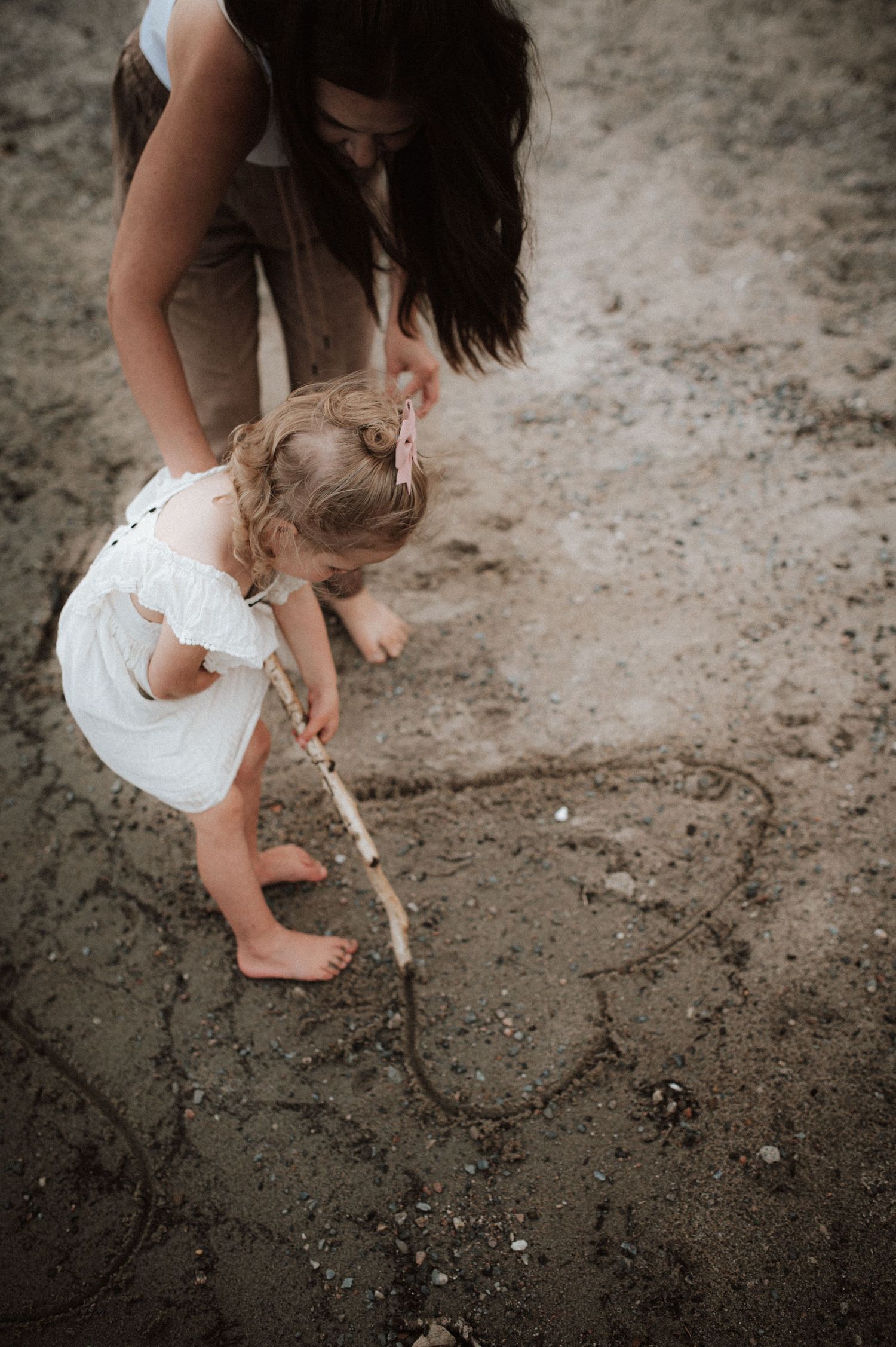 Young person in a white dress draws on sandy ground with a stick while someone guides their hand in a sepia-toned photograph.