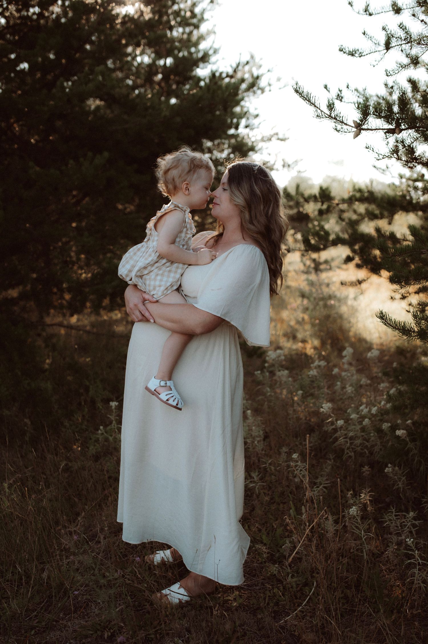 Mother in white dress holding small child while standing in nature during golden hour at sunset.