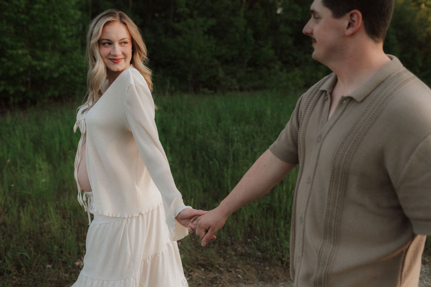 A couple in wedding attire holds hands and dances together in a grassy outdoor setting at sunset.