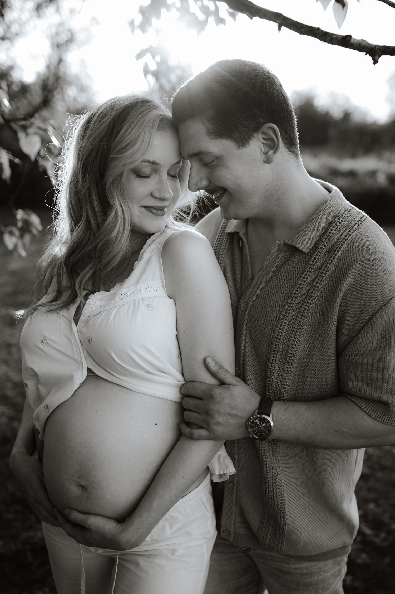 Expecting parents embrace tenderly during black and white maternity photoshoot outdoors with tree branches visible.