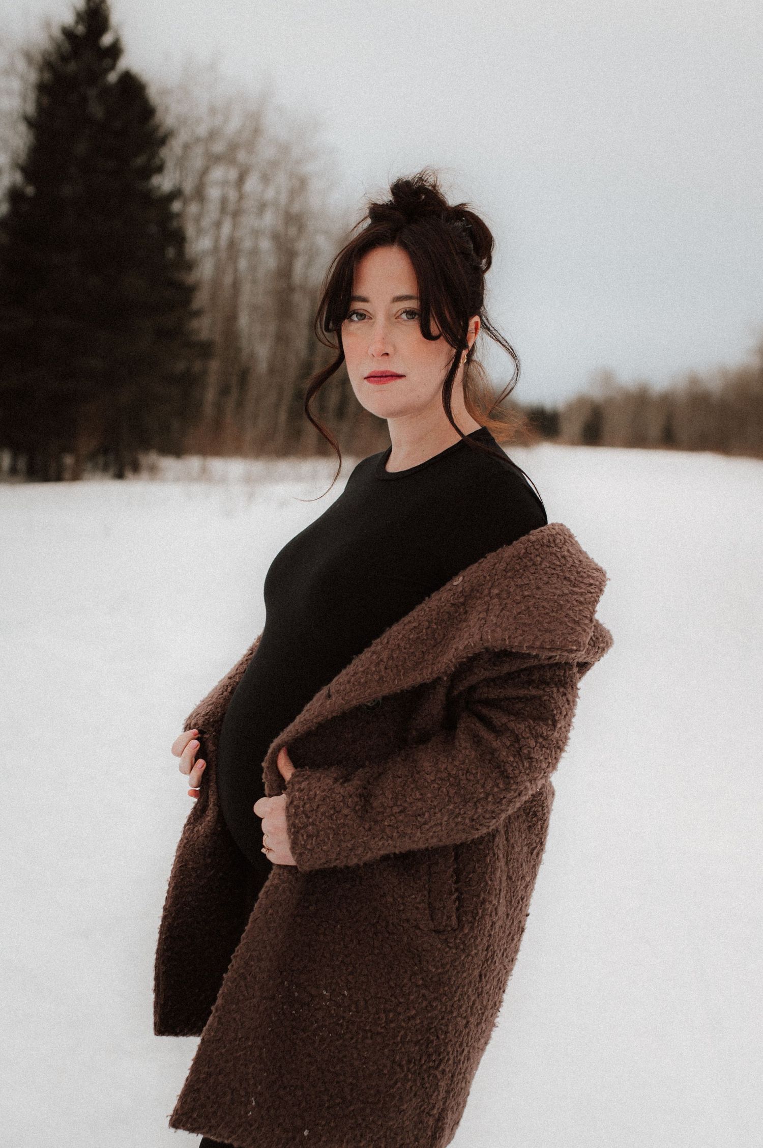 Woman wearing black sweater and brown sherpa coat poses gracefully in snowy winter landscape among evergreen trees.