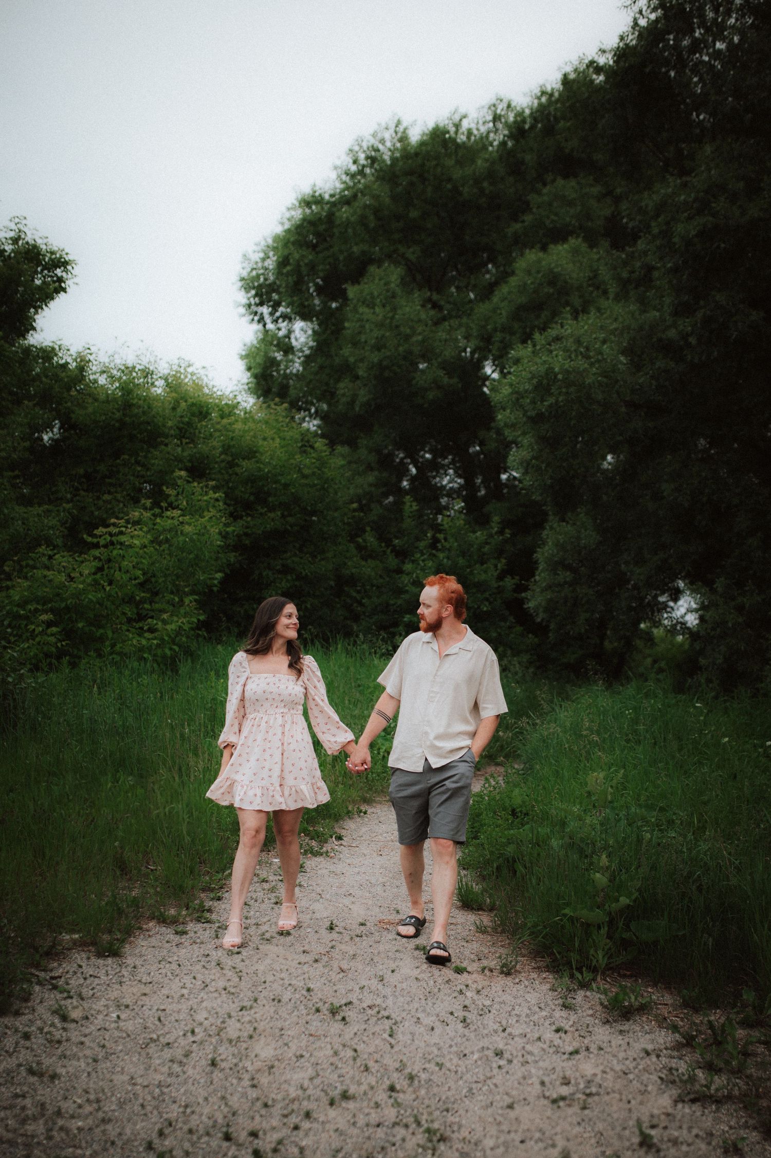A couple strolls hand in hand along a gravel path surrounded by lush green vegetation on a summer day.