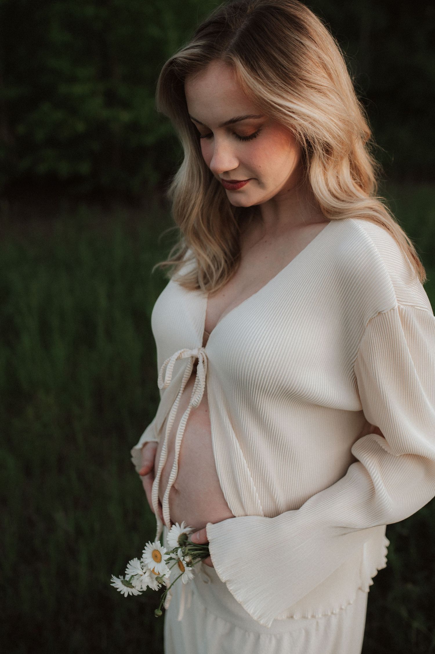 A person in a cream colored sweater holds white wildflowers while standing in a grassy outdoor setting at dusk.