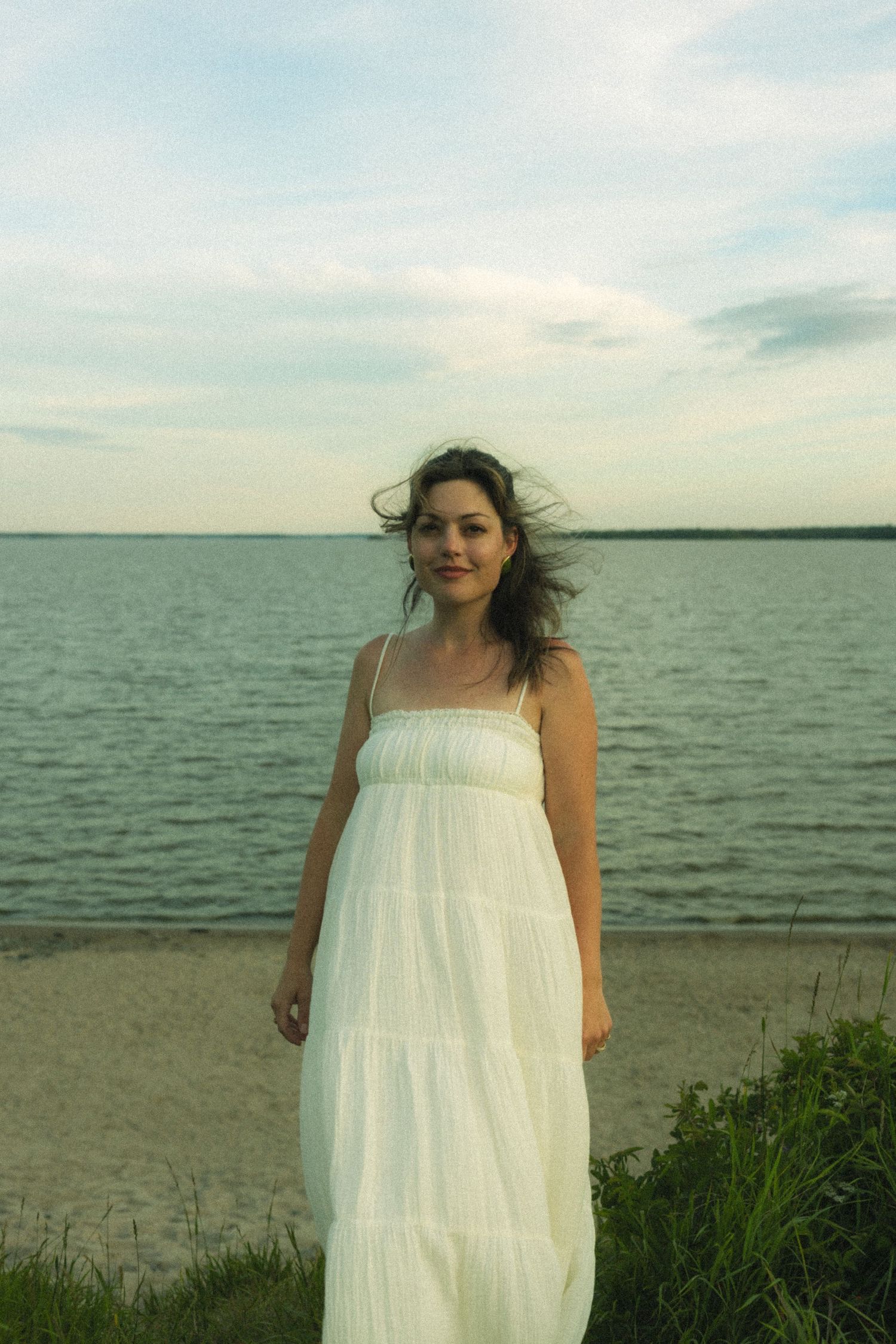 A person in a flowing white sundress stands on a beach with water and cloudy sky in the background.