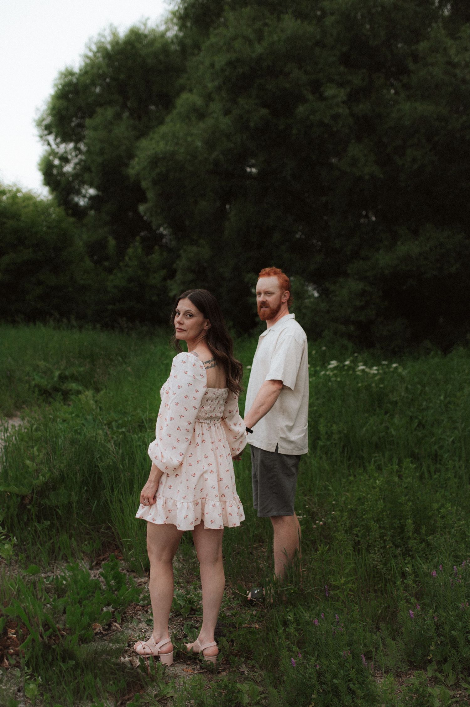 A couple stands together in a grassy field with trees in the background during golden hour.