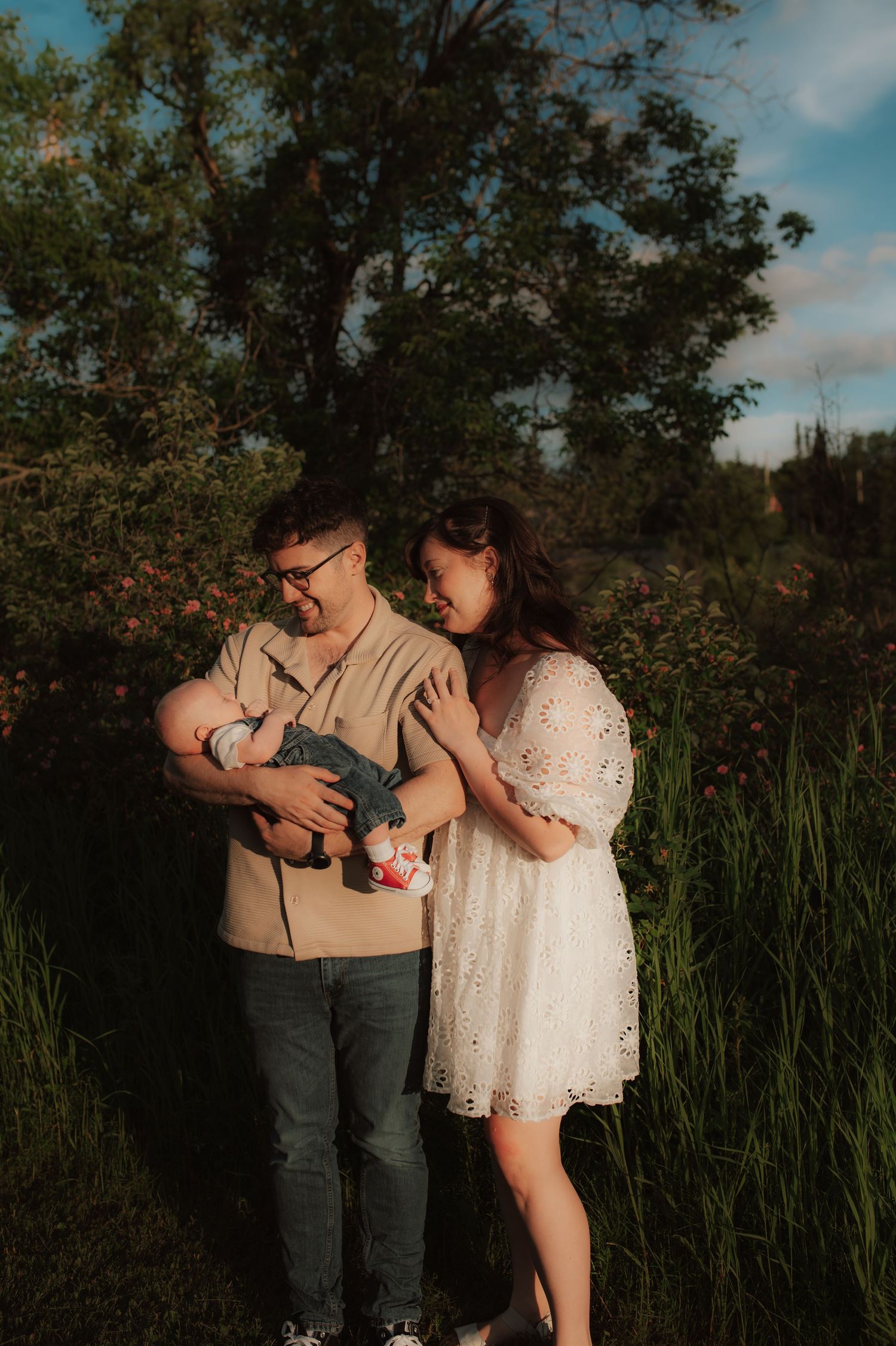 A couple in casual attire stands together outdoors during golden hour among tall grass and trees in a natural setting.