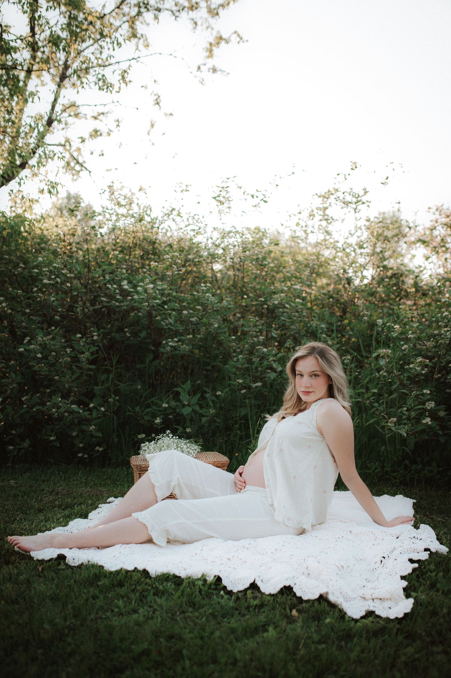 A figure in a flowing white dress reclines on grass against a backdrop of green foliage in a dreamy outdoor setting.