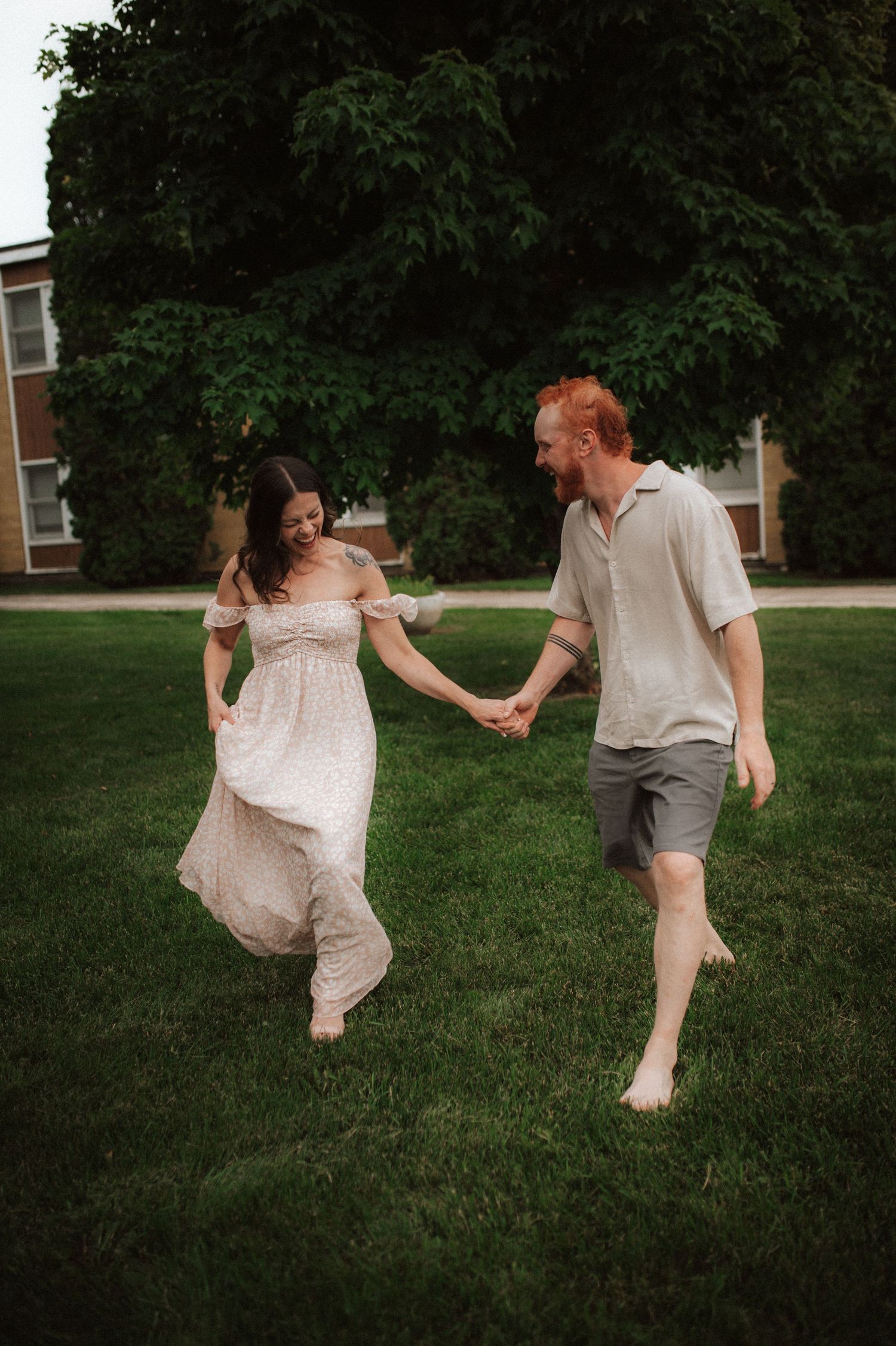 A couple runs barefoot through green grass, with her flowing white dress swaying in the summer breeze.